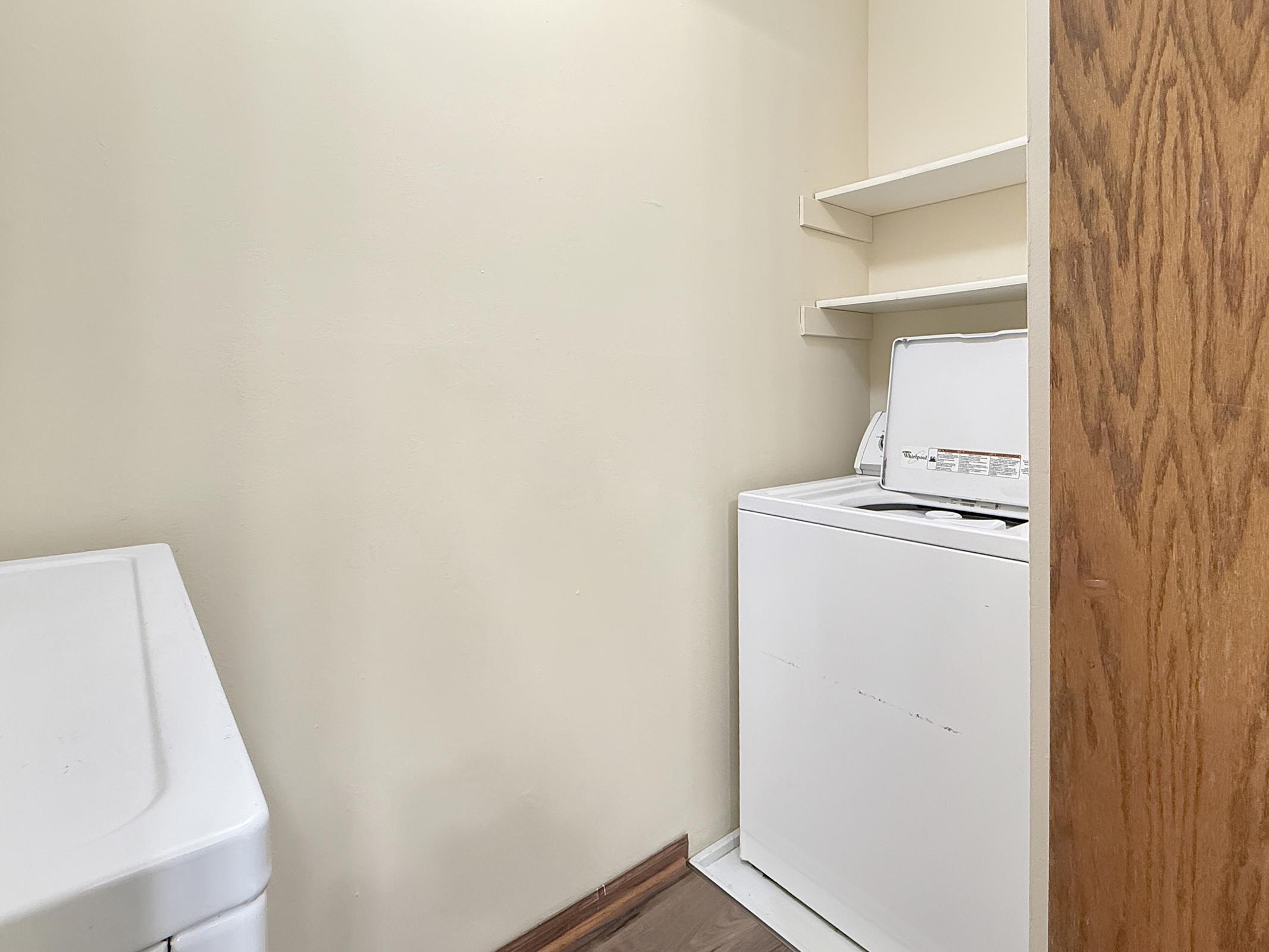 A small laundry room featuring a white washing machine and a dryer. There are shelves mounted on the wall above the appliances, and the walls are painted a soft beige color. A wooden door is visible on the right side, providing access to the room. The floor is a light-colored laminate.