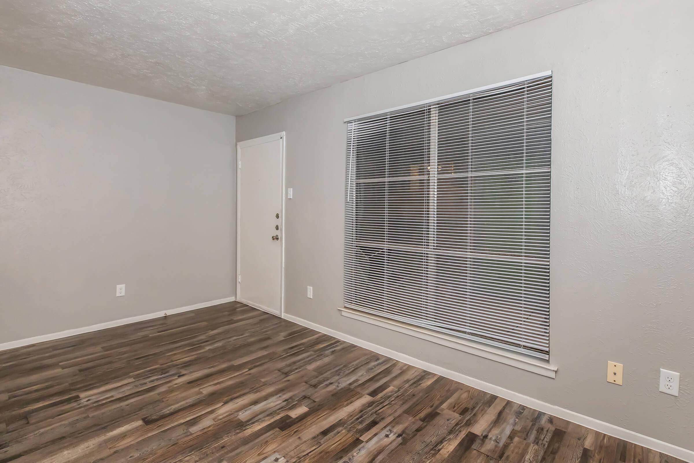 Empty interior of a room with gray walls and wood-like flooring. A closed door is on the left, and a large window with white blinds is on the right, allowing some natural light into the space. The room is unfurnished and appears to be freshly painted.