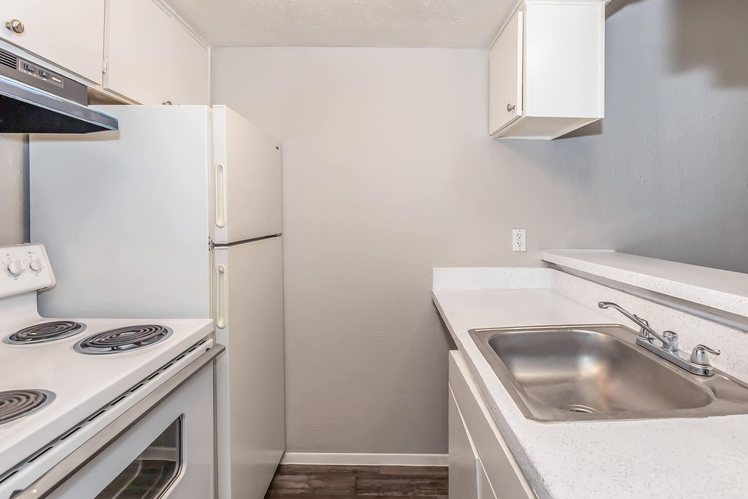 A small, modern kitchen featuring a white stove and oven, a white refrigerator, a sink with a faucet, and light-colored cabinets. The countertops are a sleek, solid surface, and the walls are painted a soft gray, creating a clean and inviting space.