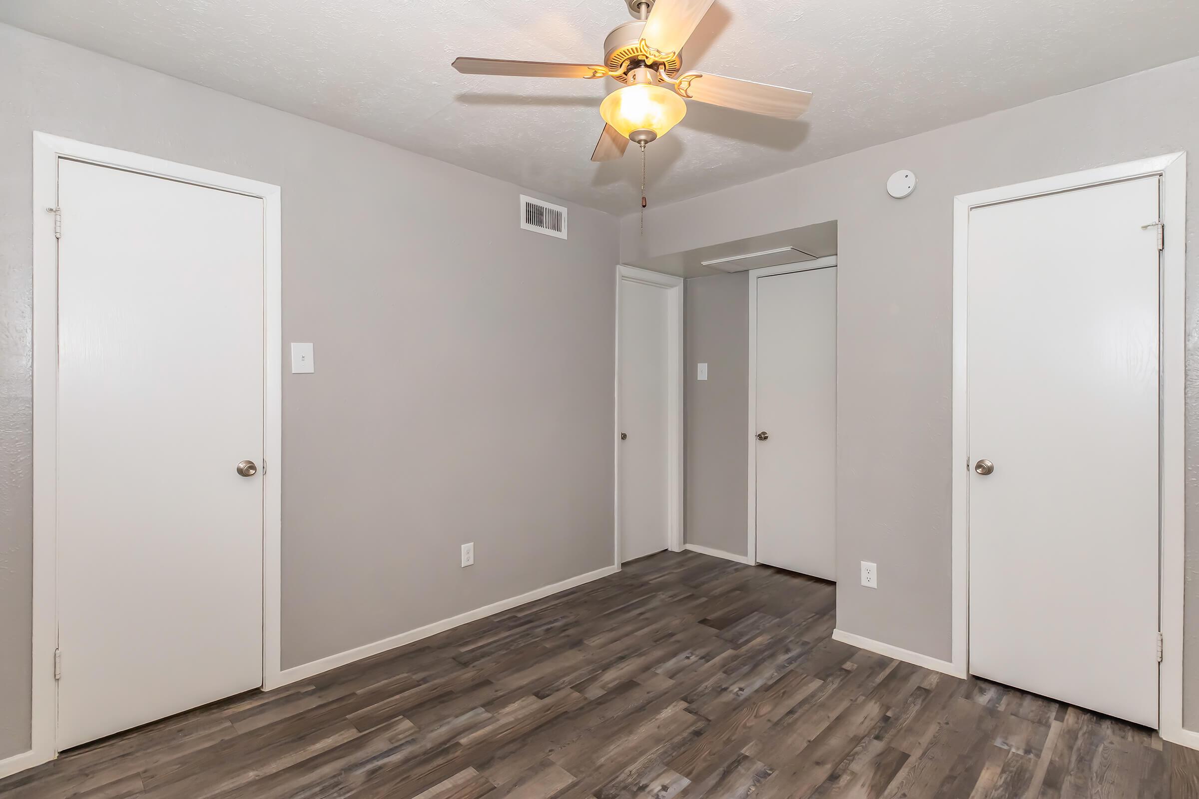 A small room with light gray walls and a ceiling fan. There are two closed doors on the left and one on the right. The floor is covered with dark wood laminate. A light fixture hangs from the ceiling, illuminating the space. The room appears empty and well-maintained.