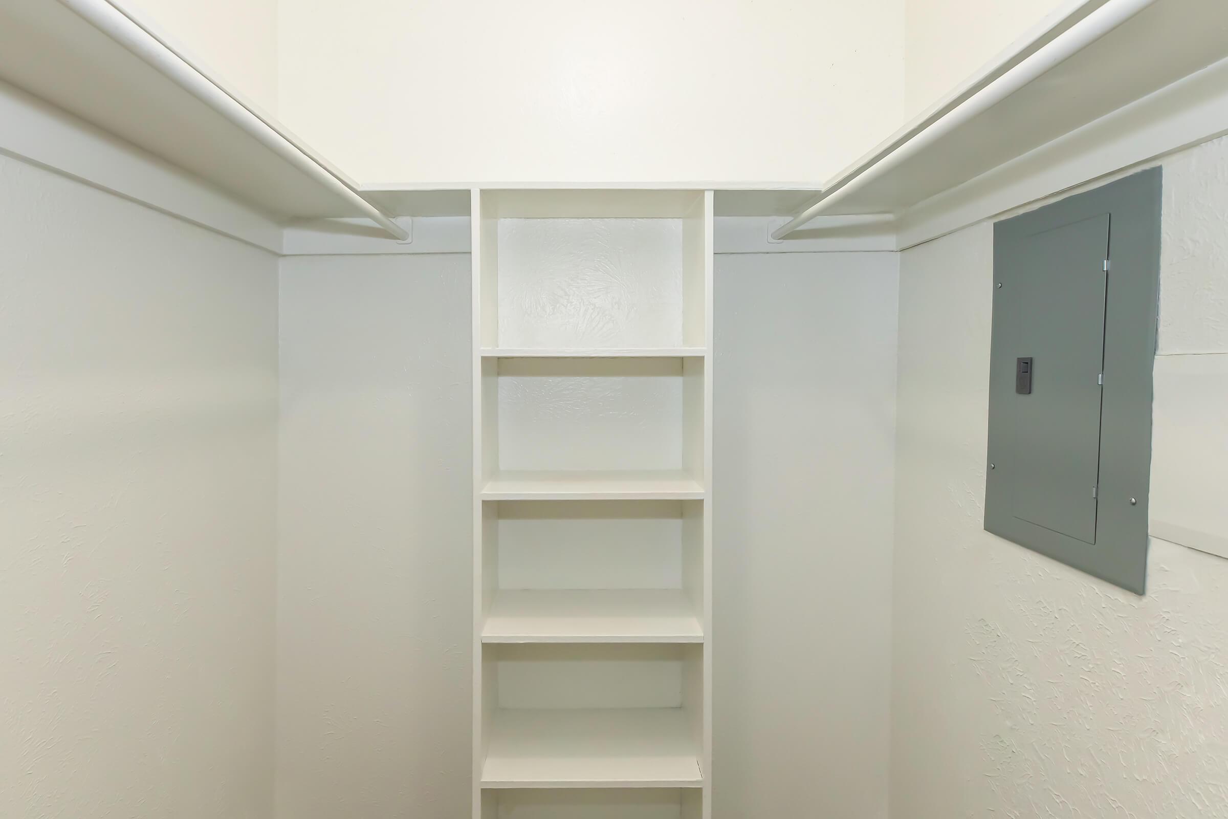 A minimalistic empty closet with white walls, featuring two hanging rods at the top and a set of five white shelves in the center. A gray electrical panel is located on the right side of the wall. The space is well-lit and has a clean, tidy appearance.