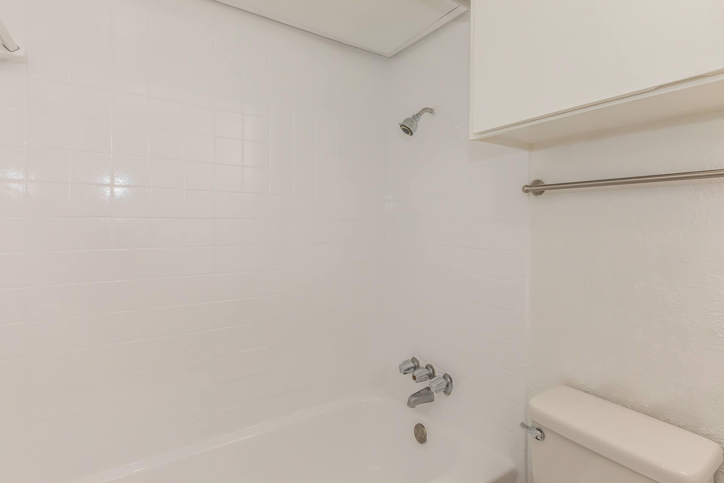 A clean, minimalist bathroom with white tiled walls, featuring a bathtub with a showerhead, a faucet, and a simple toilet. The space is well-lit and uncluttered, giving it a fresh and modern appearance. A cabinet is visible above the bathtub area.