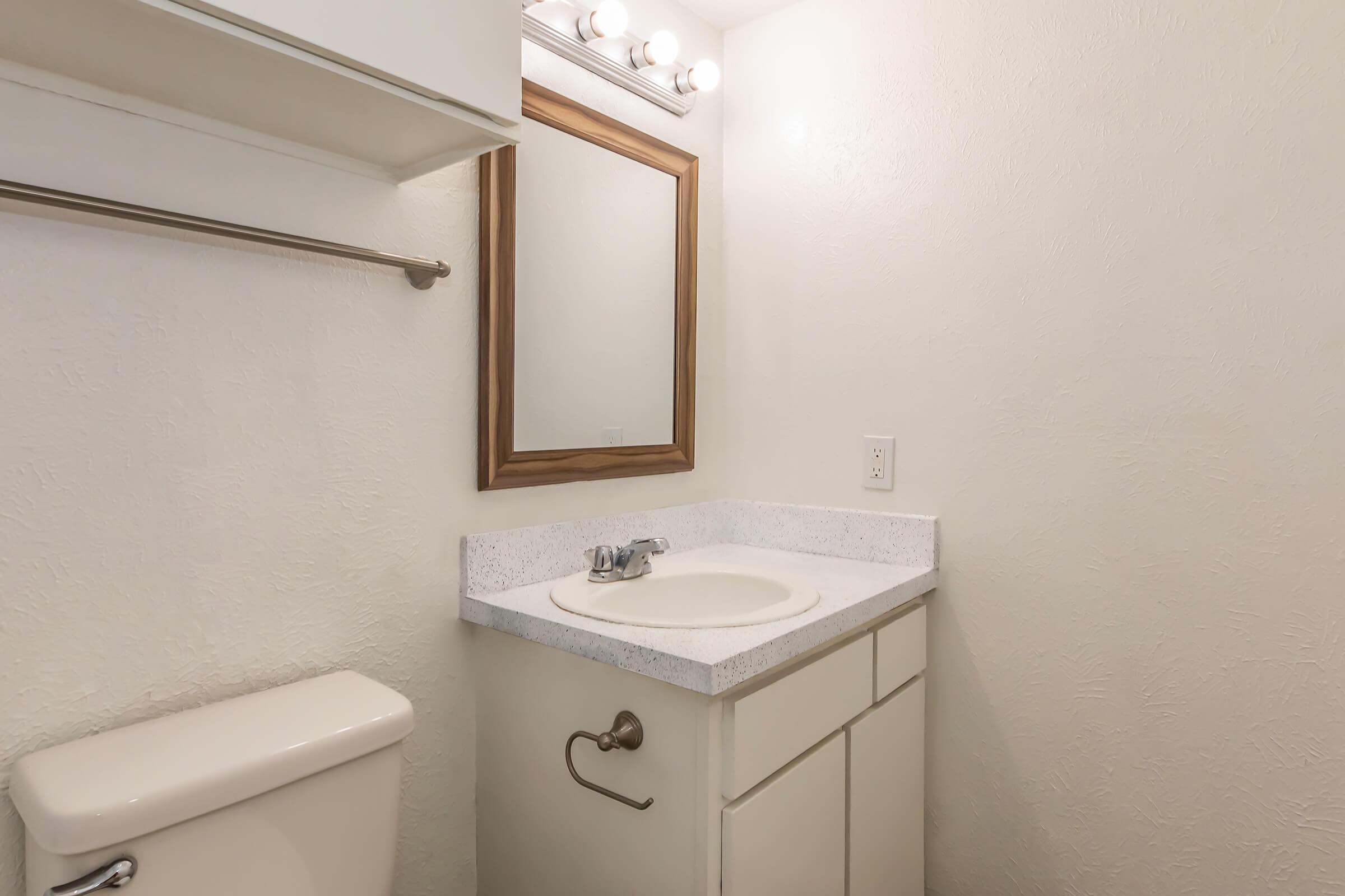 A small bathroom featuring a white sink with a countertop, a wooden-framed mirror above the sink, a toilet to the left, and a wall-mounted towel bar. The walls are painted light colors, contributing to a clean and simple aesthetic. Good lighting from the overhead fixtures enhances the space.