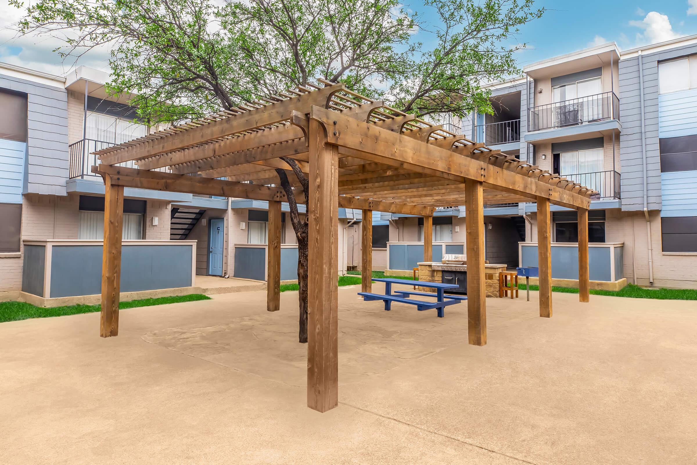 A wooden pergola with a tree in the center stands in an outdoor courtyard, surrounded by apartment buildings. Beneath the pergola, there are picnic tables with benches. The scene is bright and inviting, featuring green grass and a clear blue sky.