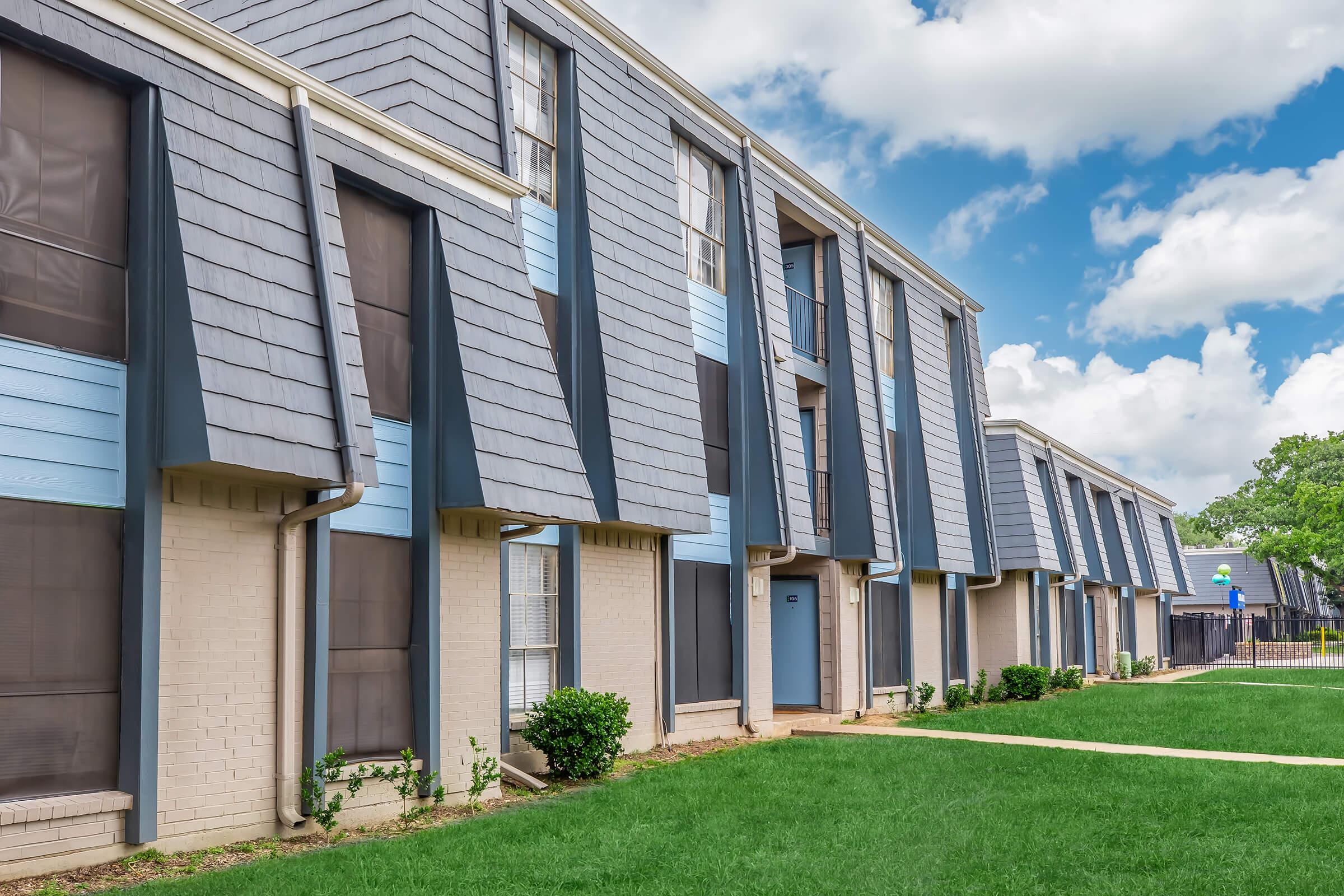 A row of modern apartment buildings with blue and gray exteriors. The buildings feature slanted roofs and large windows. A well-maintained lawn with green grass is in the foreground, and the sky is partly cloudy. The overall atmosphere is inviting and residential.