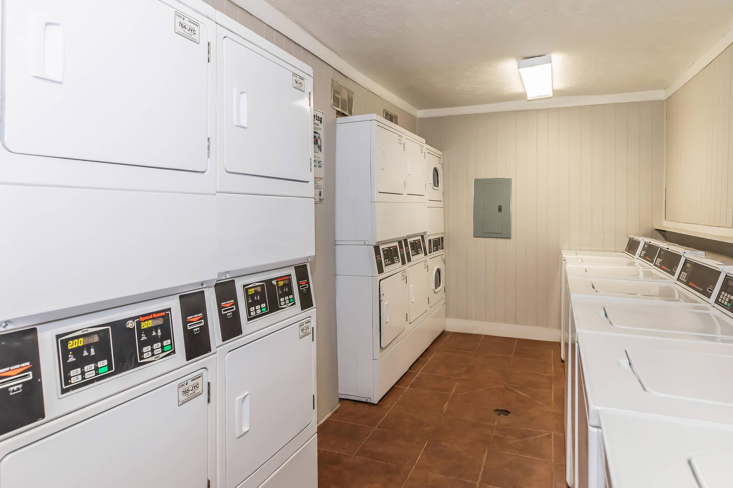 A clean and organized laundry room featuring multiple stacked washers and dryers, with clear digital displays. The walls are light-colored, and the floor has brown tiles. Bright overhead lighting enhances the spacious feel of the room.
