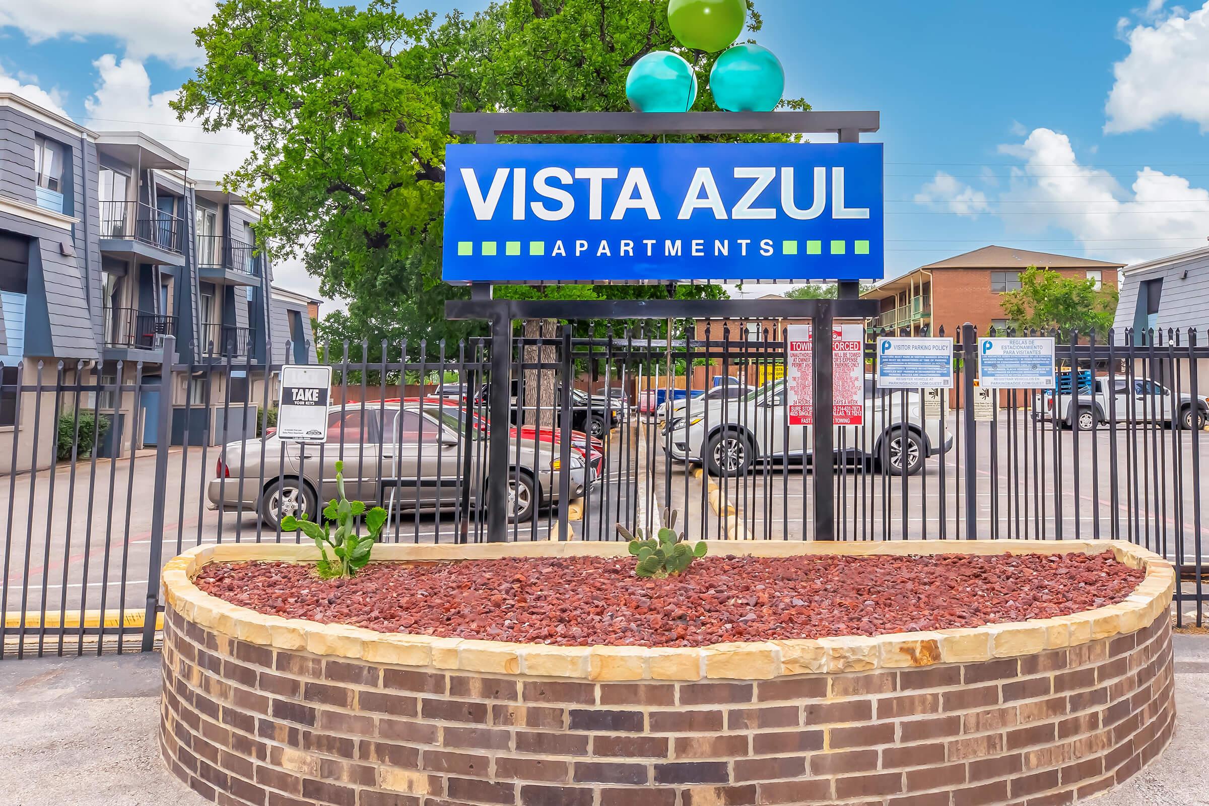 Sign for Vista Azul Apartments featuring blue and green colors, surrounded by a landscaped area with plants. In the background, there are several apartment buildings and cars parked nearby, with a clear blue sky and fluffy clouds.