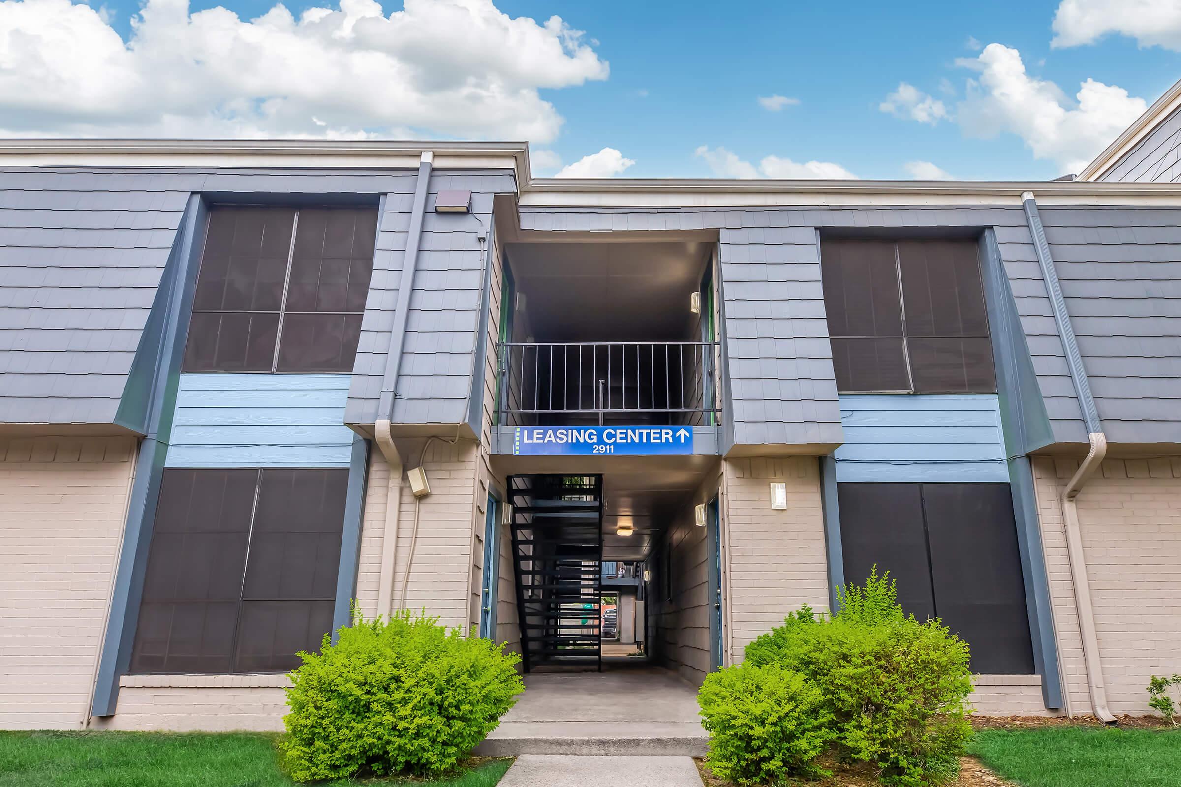Exterior view of a multi-unit residential building entrance featuring a staircase leading to a Leasing Center. The building has a gray siding design with blue accents. Lush green shrubs are positioned at the base, and there is a clear blue sky with fluffy clouds in the background.