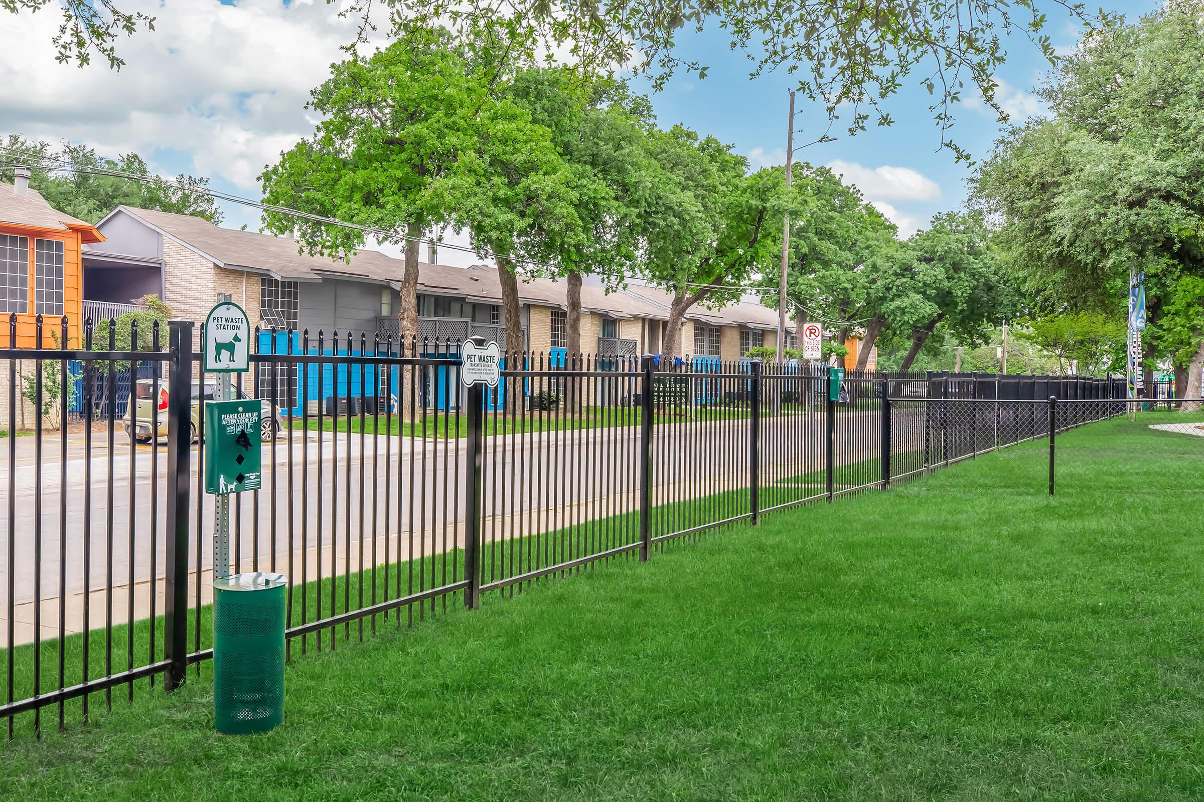 A well-maintained grassy area beside a black metal fence, with green dog waste stations and signs. In the background, residential buildings with tree coverage are visible under a partly cloudy sky, showcasing a neat, suburban neighborhood setting.