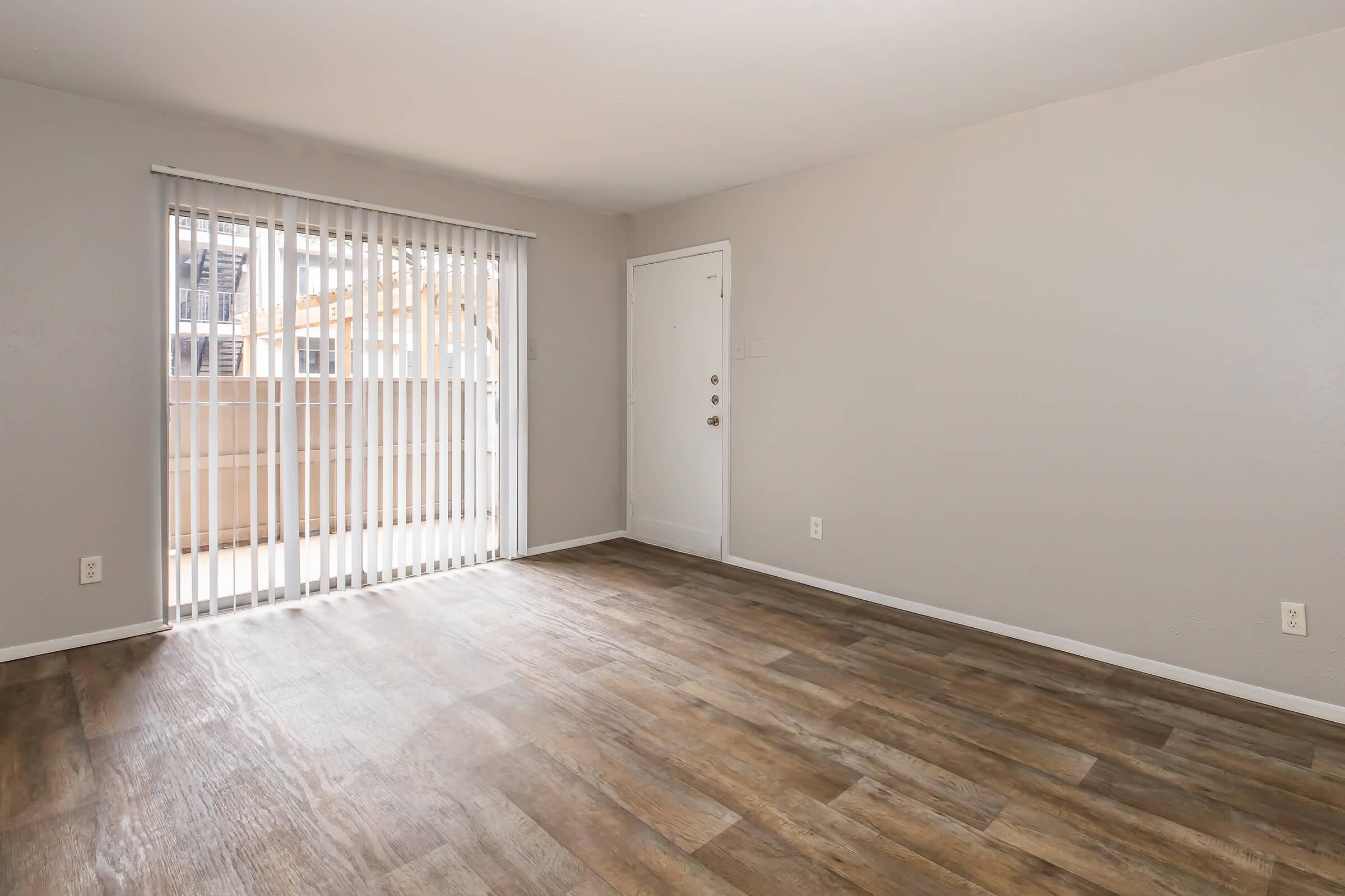 Empty room with light-colored walls and wooden flooring. A door with vertical blinds leads to a balcony. The space is well-lit and minimalistic, ideal for personalizing or decorating.