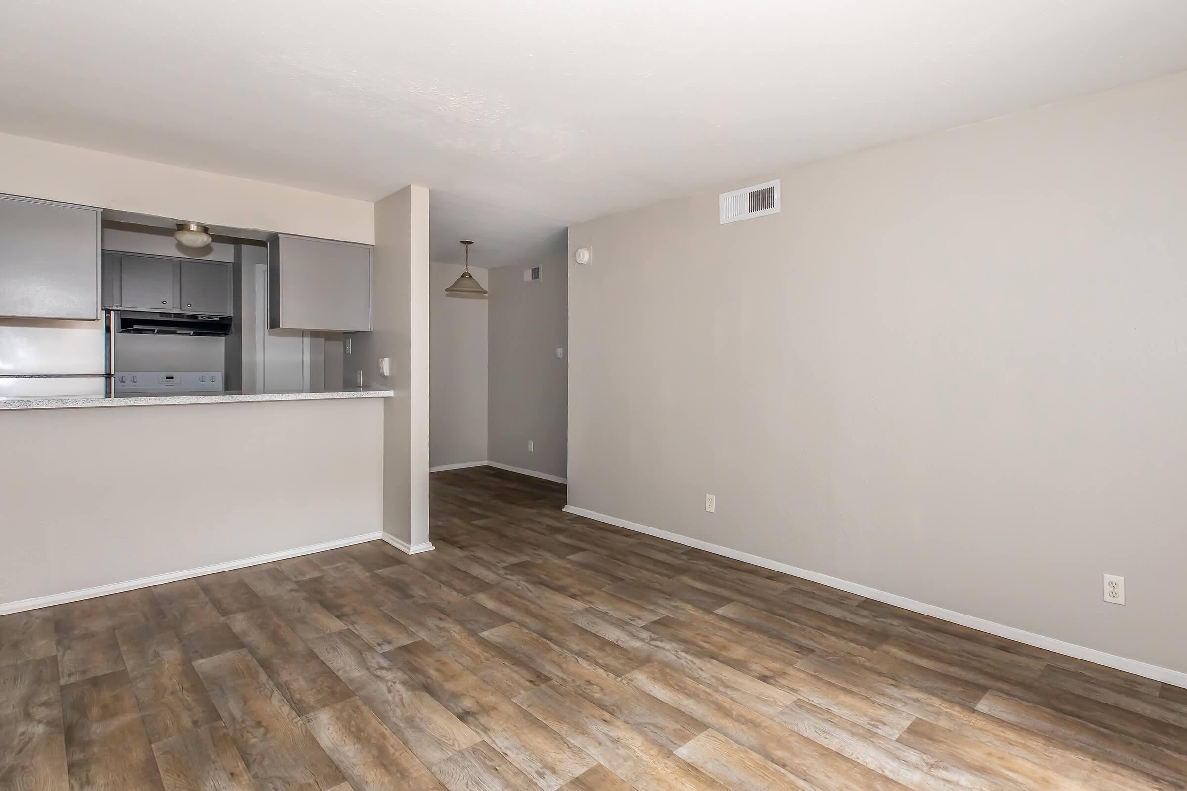 Spacious living area with light-colored walls and wood-style flooring. An open kitchen is visible in the background, featuring modern cabinets and appliances. The room has ample natural light and a neutral color scheme, creating a warm and inviting atmosphere.