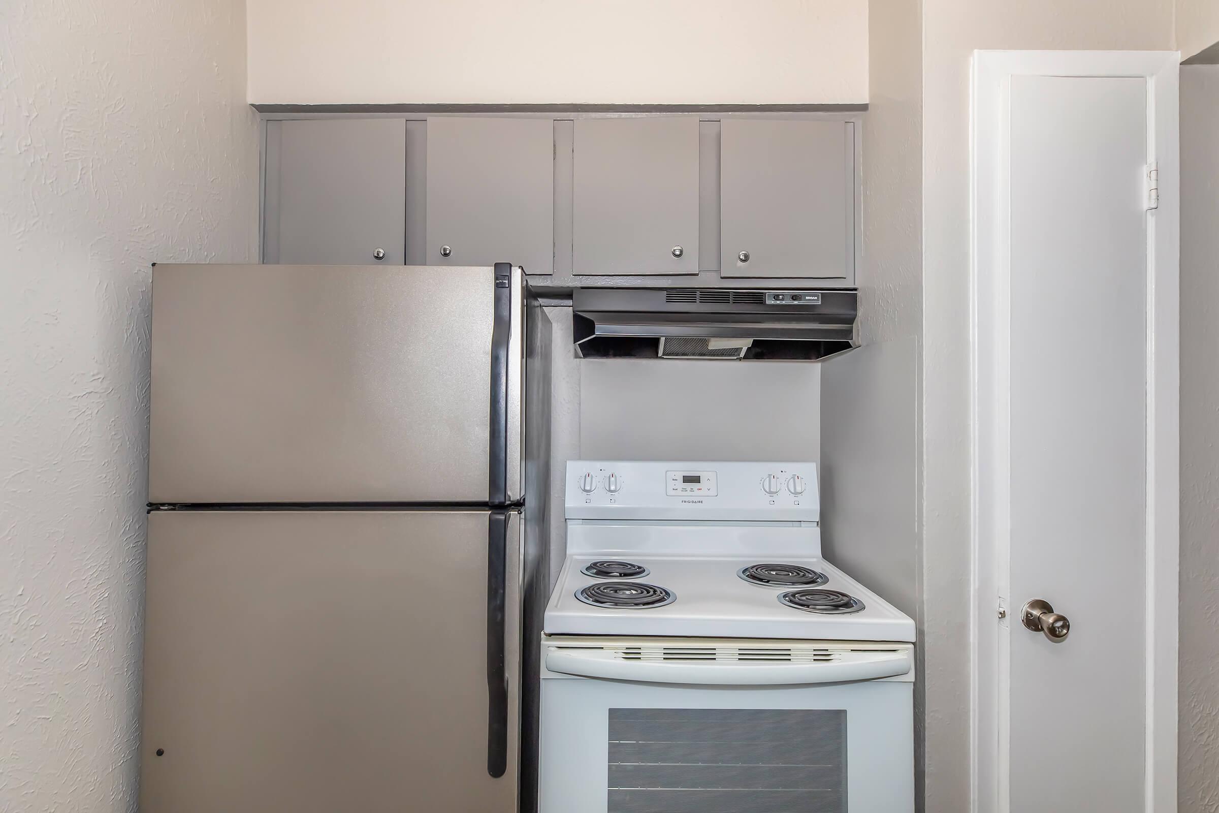 A small kitchen featuring a gray refrigerator on the left, a white stove with an oven in the center, and overhead gray cabinets. The wall is painted in a light color, and there's a white door on the right. The overall space appears clean and well-organized.