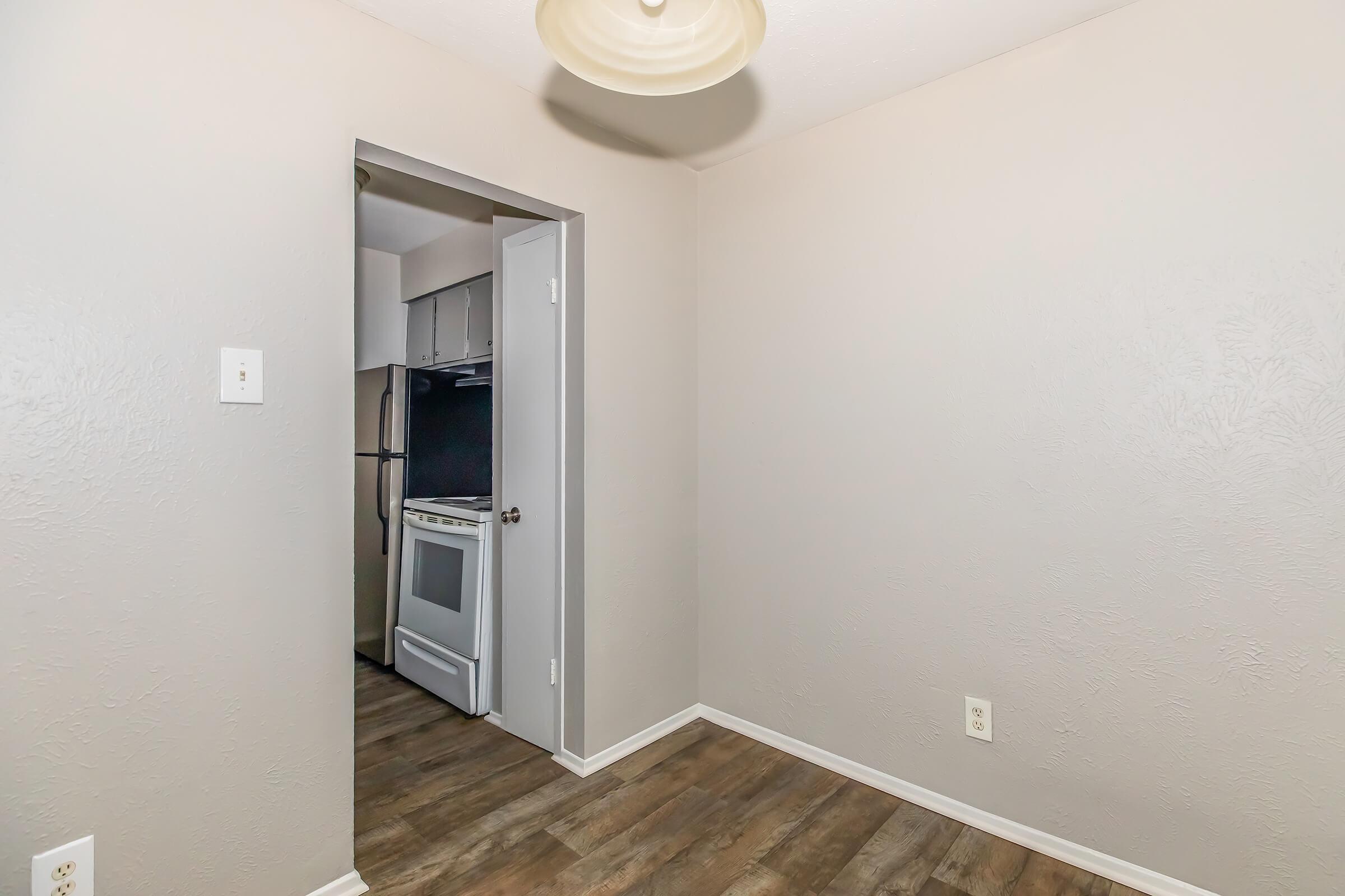 Empty room with light gray walls and a wooden floor. A doorway leads to a kitchen area featuring a visible white stove and black refrigerator. The ceiling has a simple light fixture. There are no furnishings or decorations in the visible space.