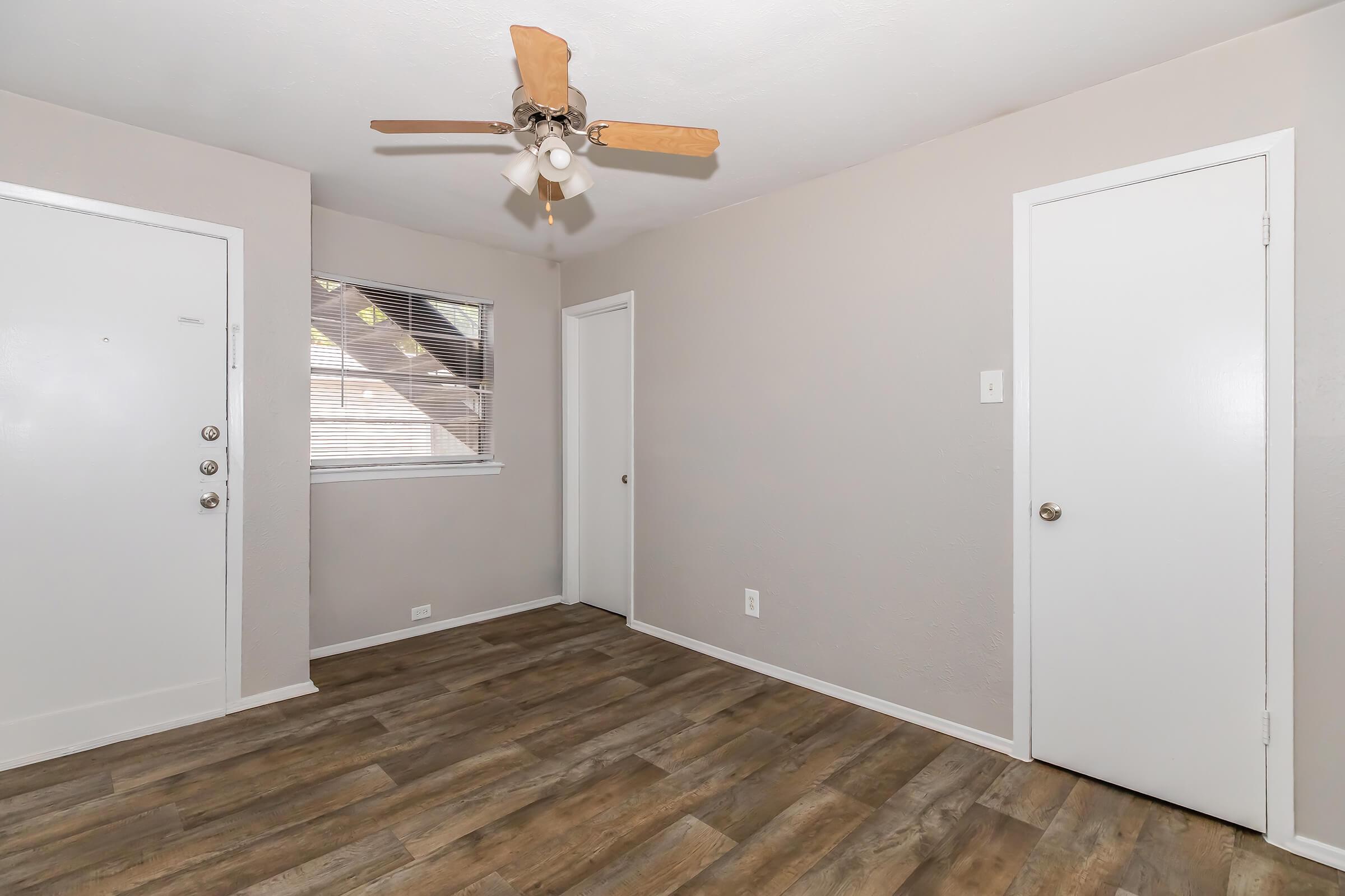A small, unfurnished room with a light brown wooden floor, a ceiling fan, and two white doors. One door leads outside, and the other opens to another space. A window with blinds allows natural light to enter. The walls are painted in a light gray color, creating a neutral and open atmosphere.