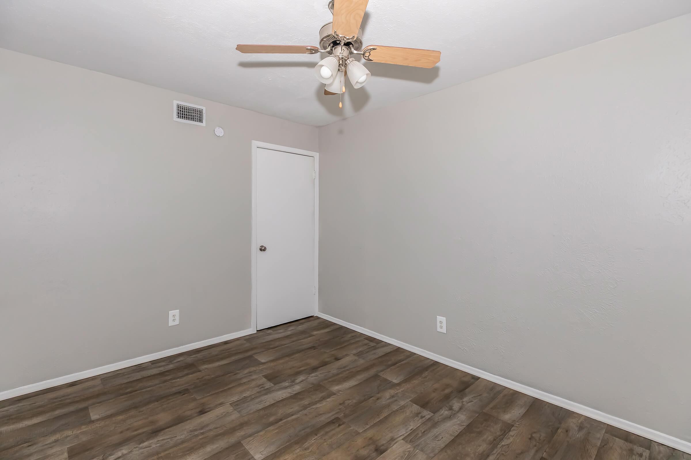 Empty room with light gray walls, wooden flooring, and a ceiling fan with four bulbs. There's a closed white door on the right and a vent on the wall. The space appears well-lit and ready for furnishing.