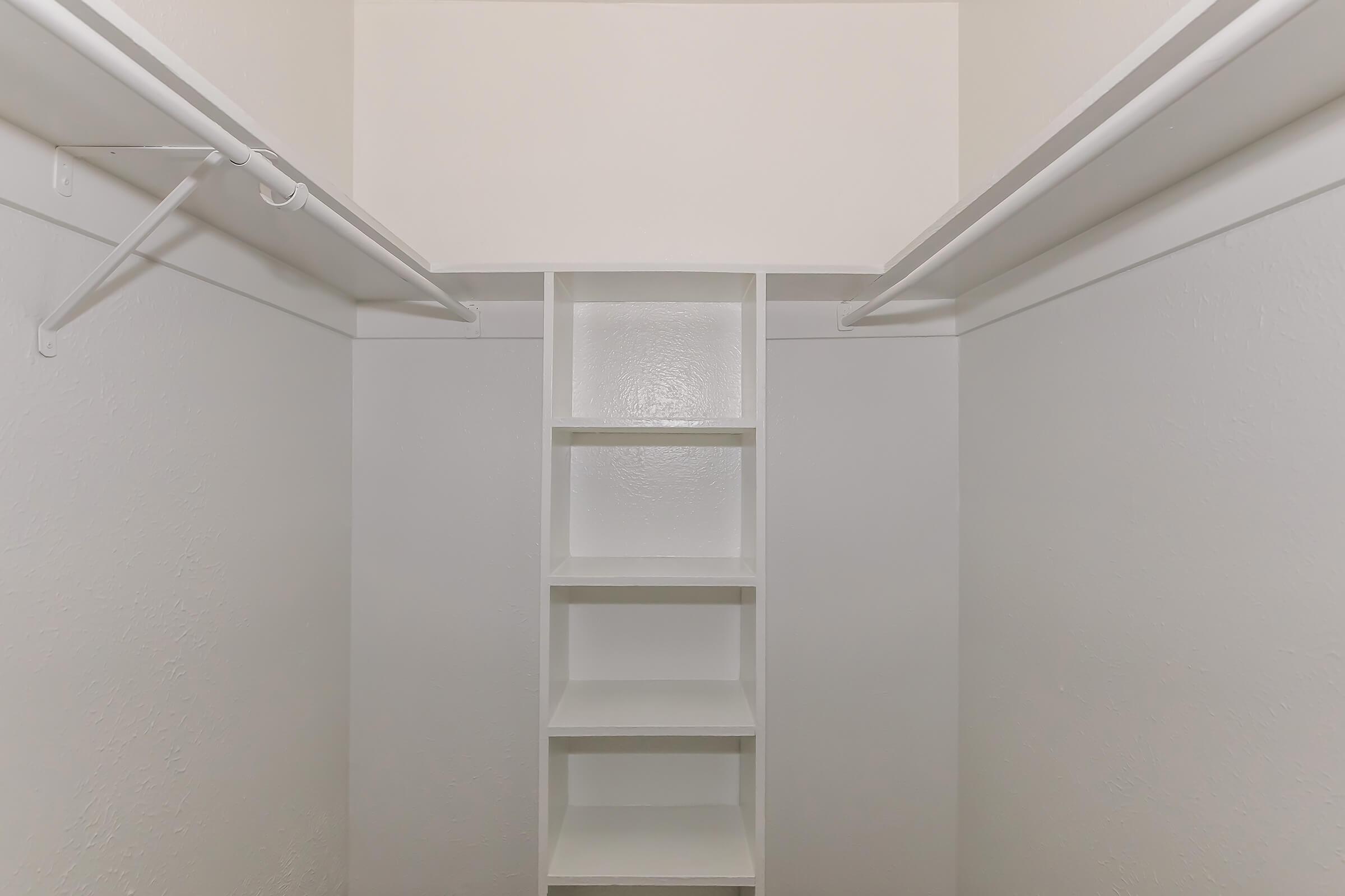 Interior of a spacious, empty closet featuring white walls, a built-in shelving unit in the center, and two hanging rods on either side. The shelves are unobstructed and provide ample storage space, creating a clean and minimalist look. The design emphasizes simplicity and organization.
