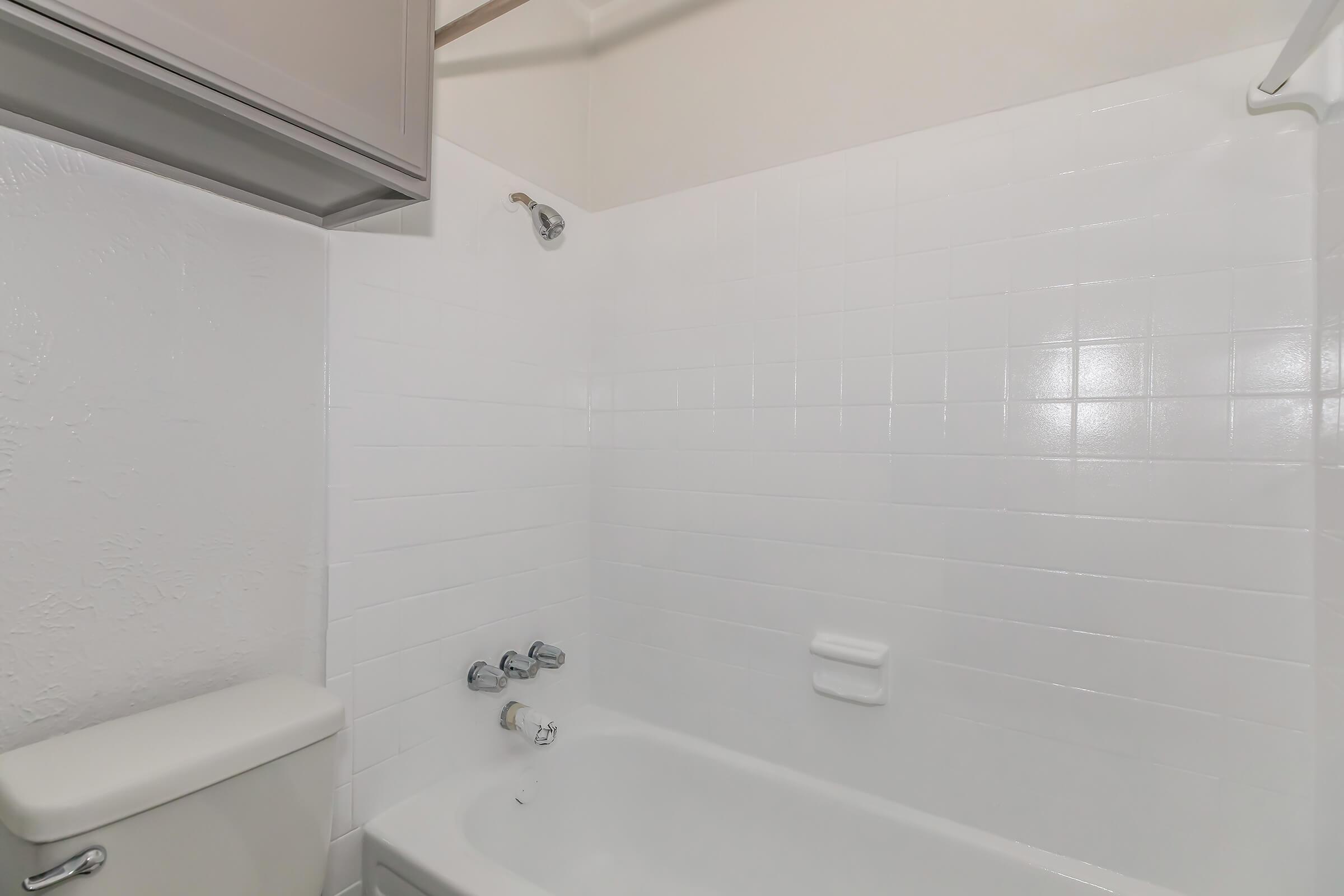 A clean, white-tiled bathroom featuring a bathtub with a showerhead above, a toilet beside it, and minimalistic walls. The overall design is simple and modern, with bright lighting reflecting off the tiles.