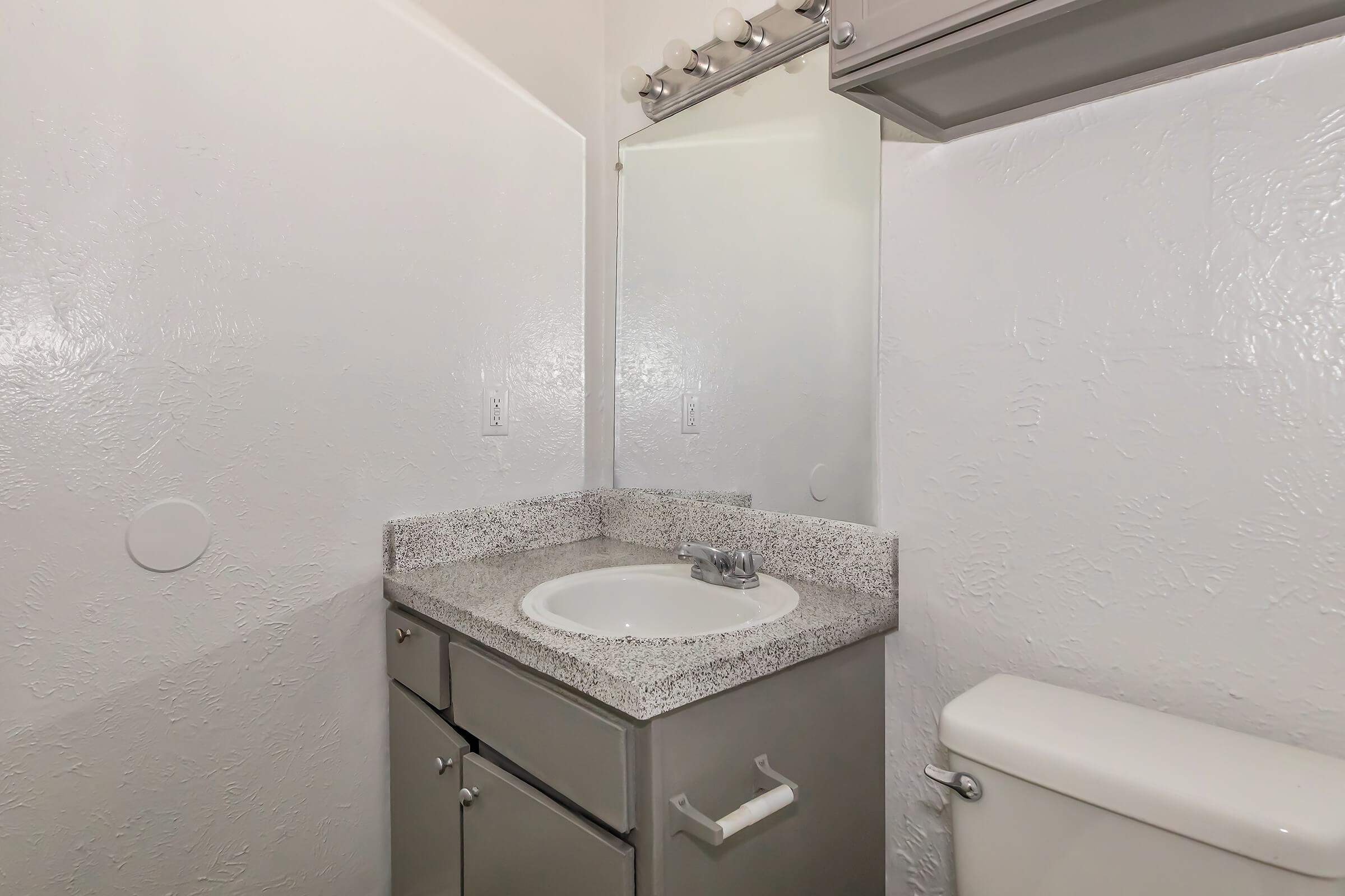 A small bathroom featuring a light-colored textured wall, a granite countertop with a sink, a mirror above the sink, and a toilet. The design is simple and modern, with minimal decorations. Bright lighting emphasizes the clean appearance of the space.