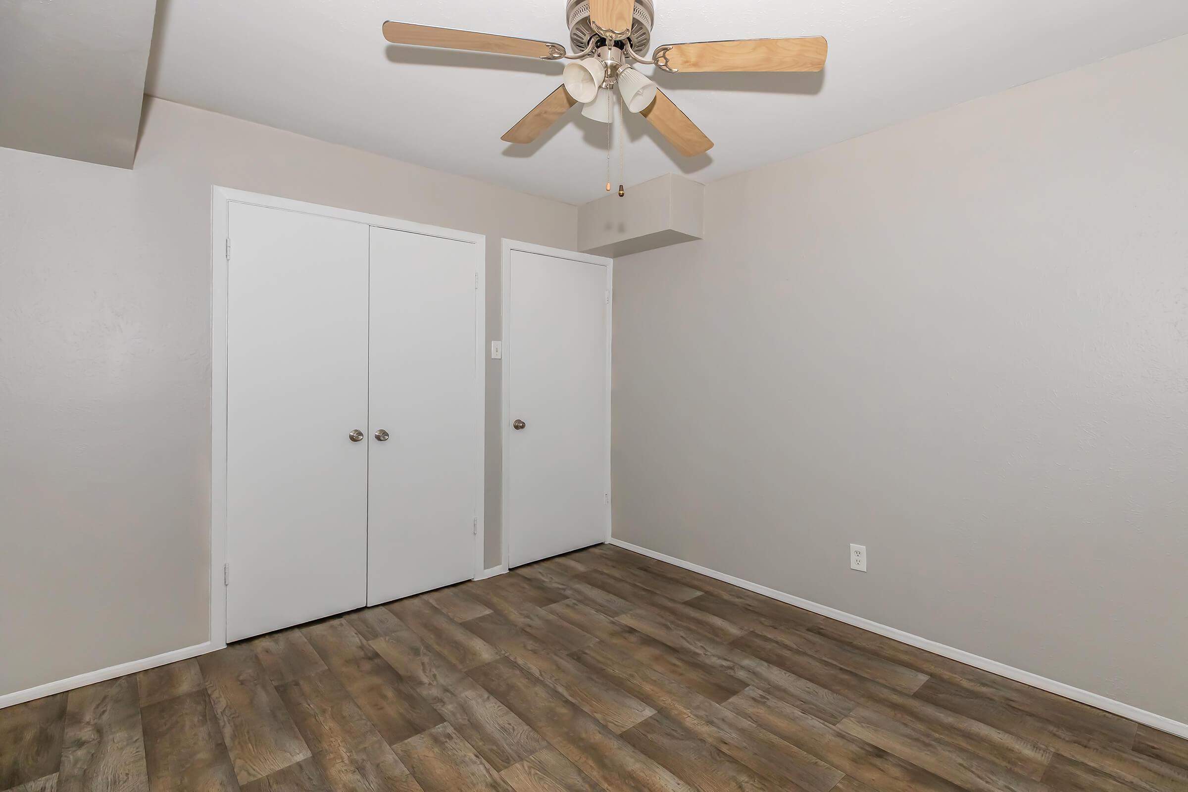 Empty room featuring light-colored walls, two closed white doors on the left, and a ceiling fan with wooden blades. The floor is covered in laminate wood, giving a warm appearance. No furniture or decor is present, creating a spacious and minimalist feel.