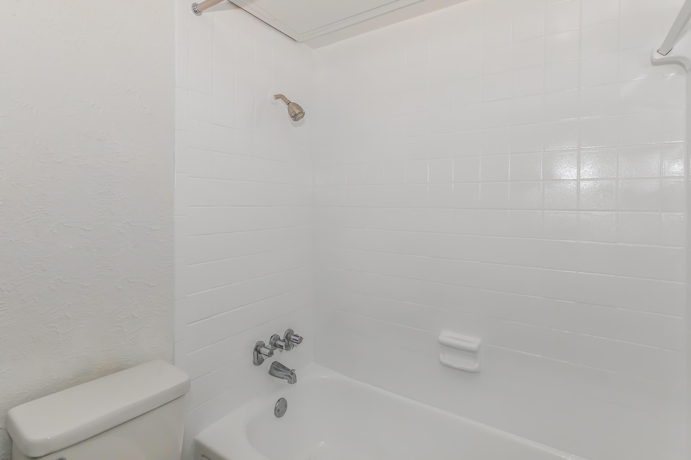 A clean and simple bathroom shower area featuring white tiled walls, a tub, a shower head, and faucet controls. The scene includes a toilet positioned beside the tub, all presented in a well-lit environment.