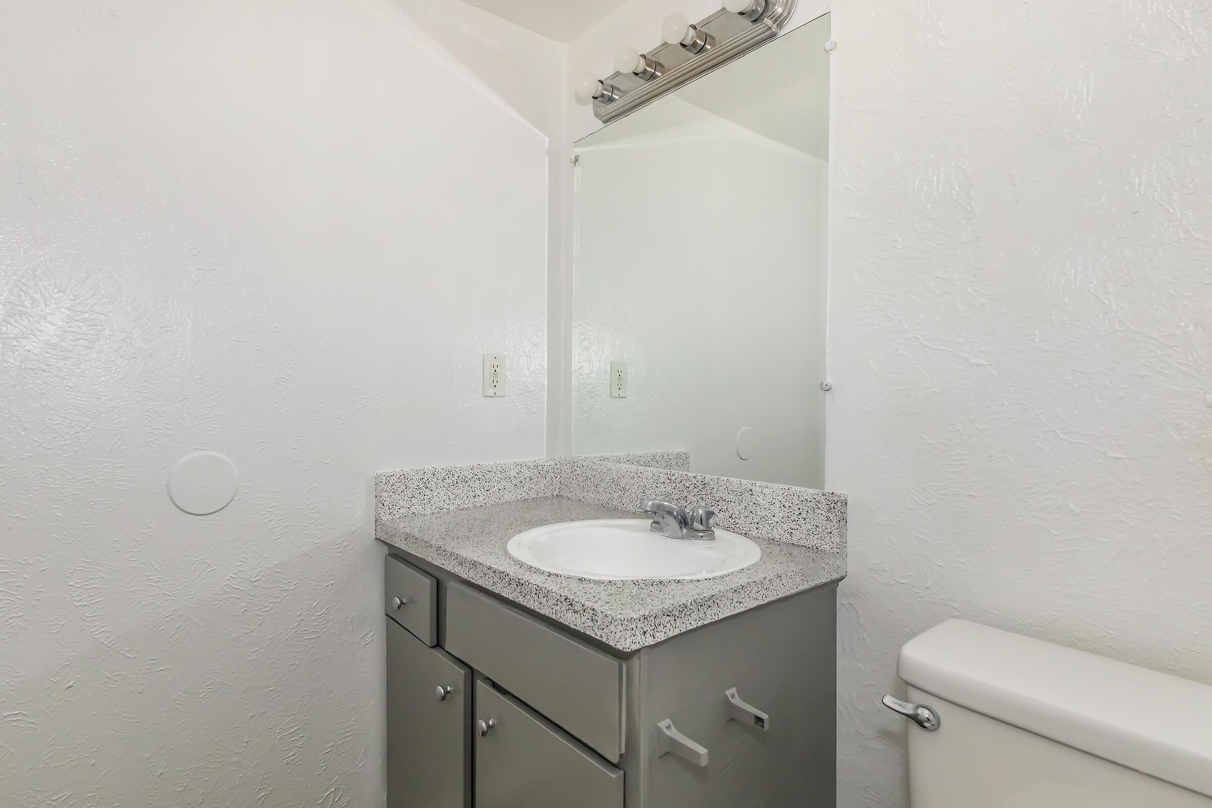 A clean bathroom featuring a gray speckled countertop with a sink, a silver faucet, a large mirror above, and a toilet. The walls are textured and white, enhancing the bright and airy feel of the space.