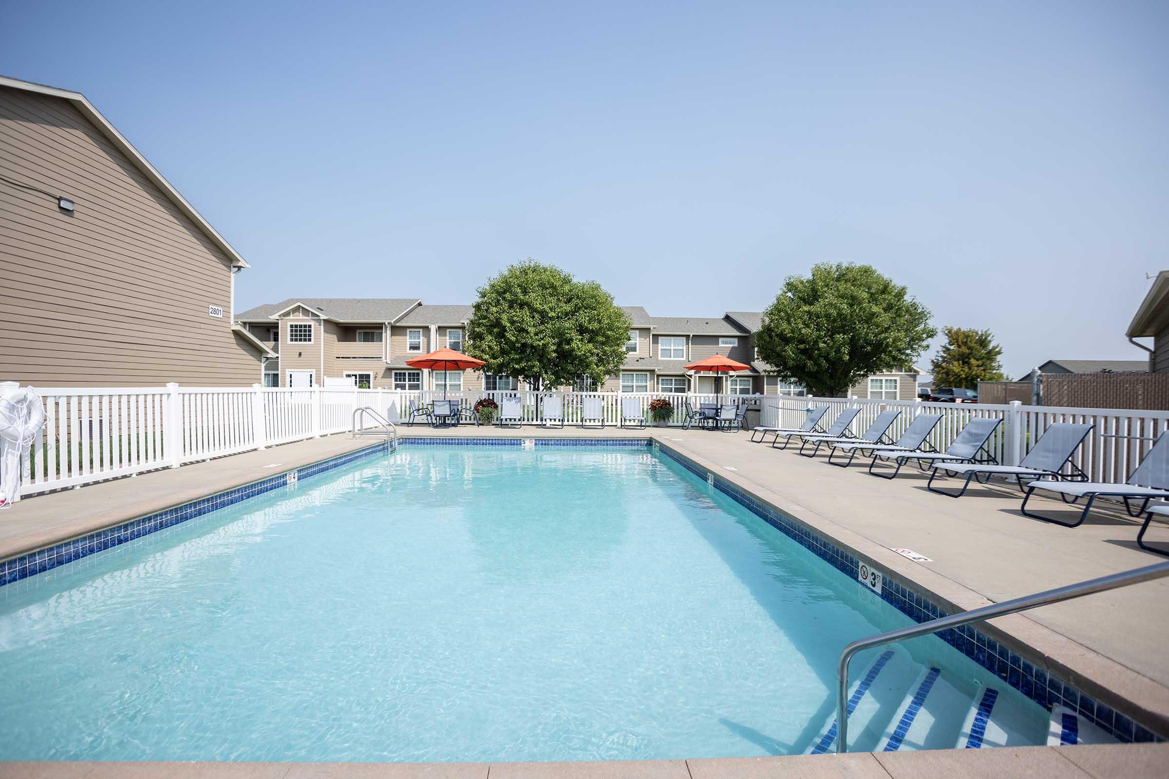 A clear swimming pool surrounded by lounge chairs, with a white fence in the foreground. In the background, there are trees and several apartment buildings. Bright blue sky overhead, creating a serene and inviting atmosphere for relaxation and leisure.