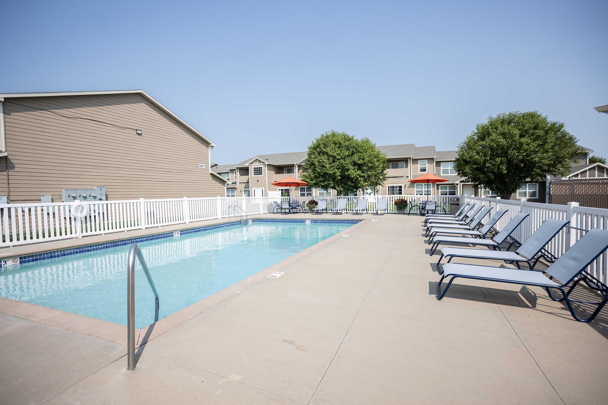 A sunny outdoor pool area featuring a clear blue swimming pool surrounded by lounge chairs. In the background, there are residential buildings with green trees and patio umbrellas offering shade. The overall atmosphere is inviting and relaxed, ideal for leisure activities.
