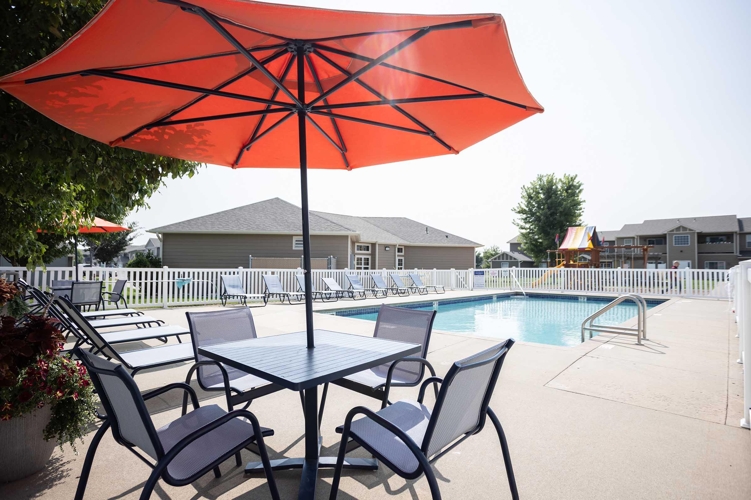 A sunny outdoor pool area with a clear blue pool surrounded by lounge chairs. A table with four chairs is shaded by a large orange umbrella. In the background, there are residential buildings and a colorful play structure. The atmosphere is inviting and perfect for relaxation.