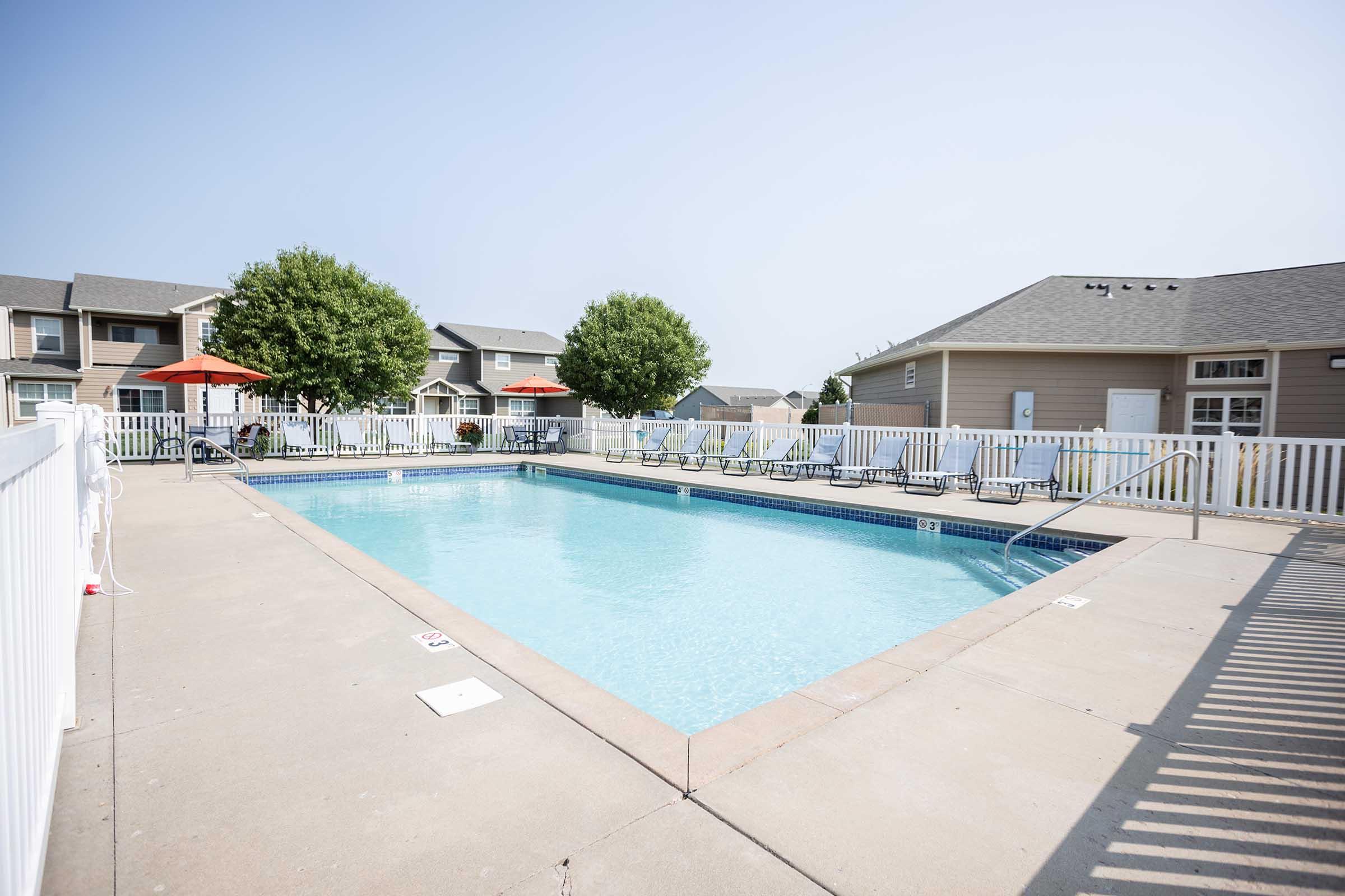 A clean swimming pool surrounded by lounge chairs and umbrellas, with a light-colored deck and white fencing, set in a residential area with apartment buildings in the background and clear blue skies.