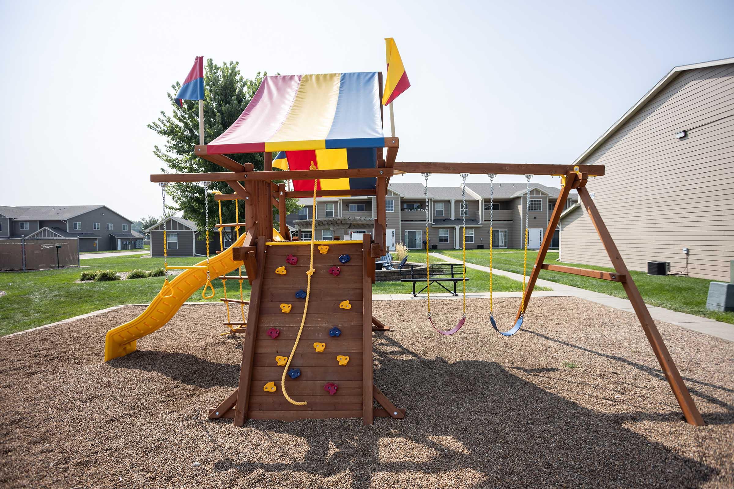 A colorful wooden playground structure featuring a yellow slide, climbing wall with colorful grips, swings, and a canopy, situated on a gravel surface. In the background, there are several apartment buildings and a grassy area.