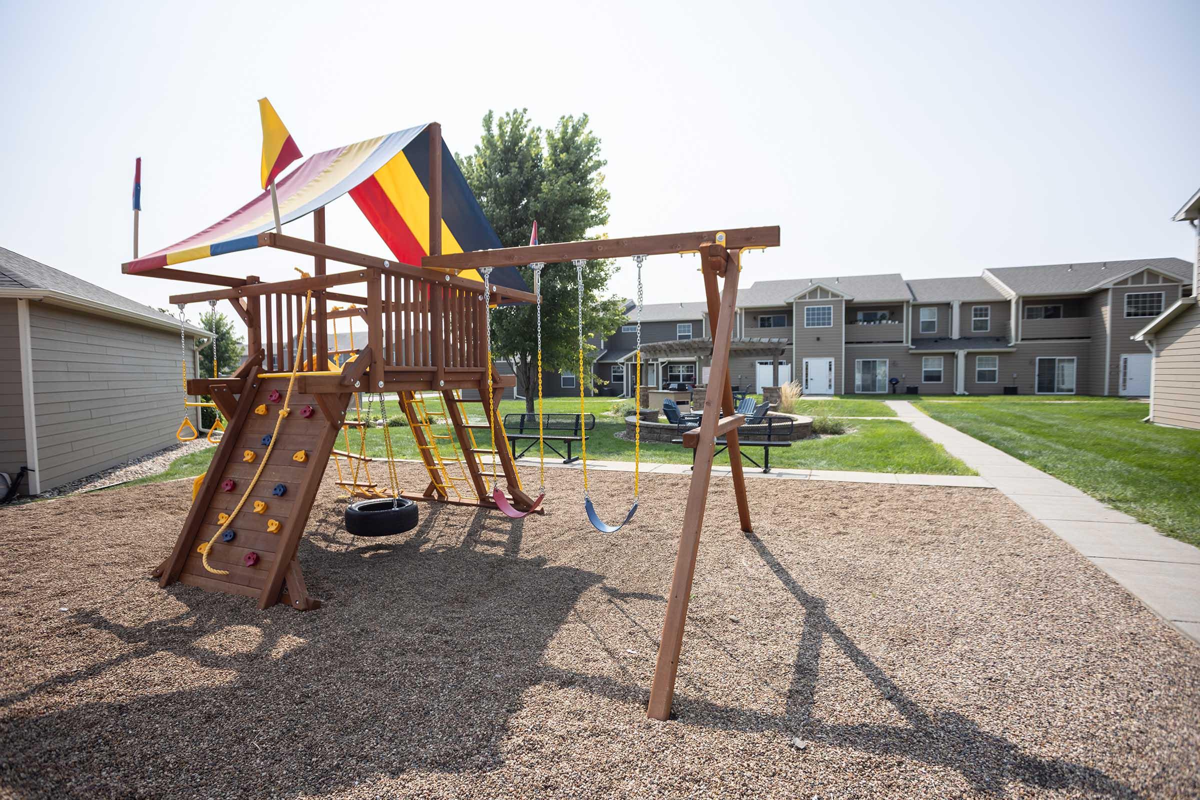 A colorful wooden playset with a slide, climbing wall, swings, and a tire swing in a gravel area, surrounded by green grass and trees. Apartment buildings are visible in the background, under a clear sky.