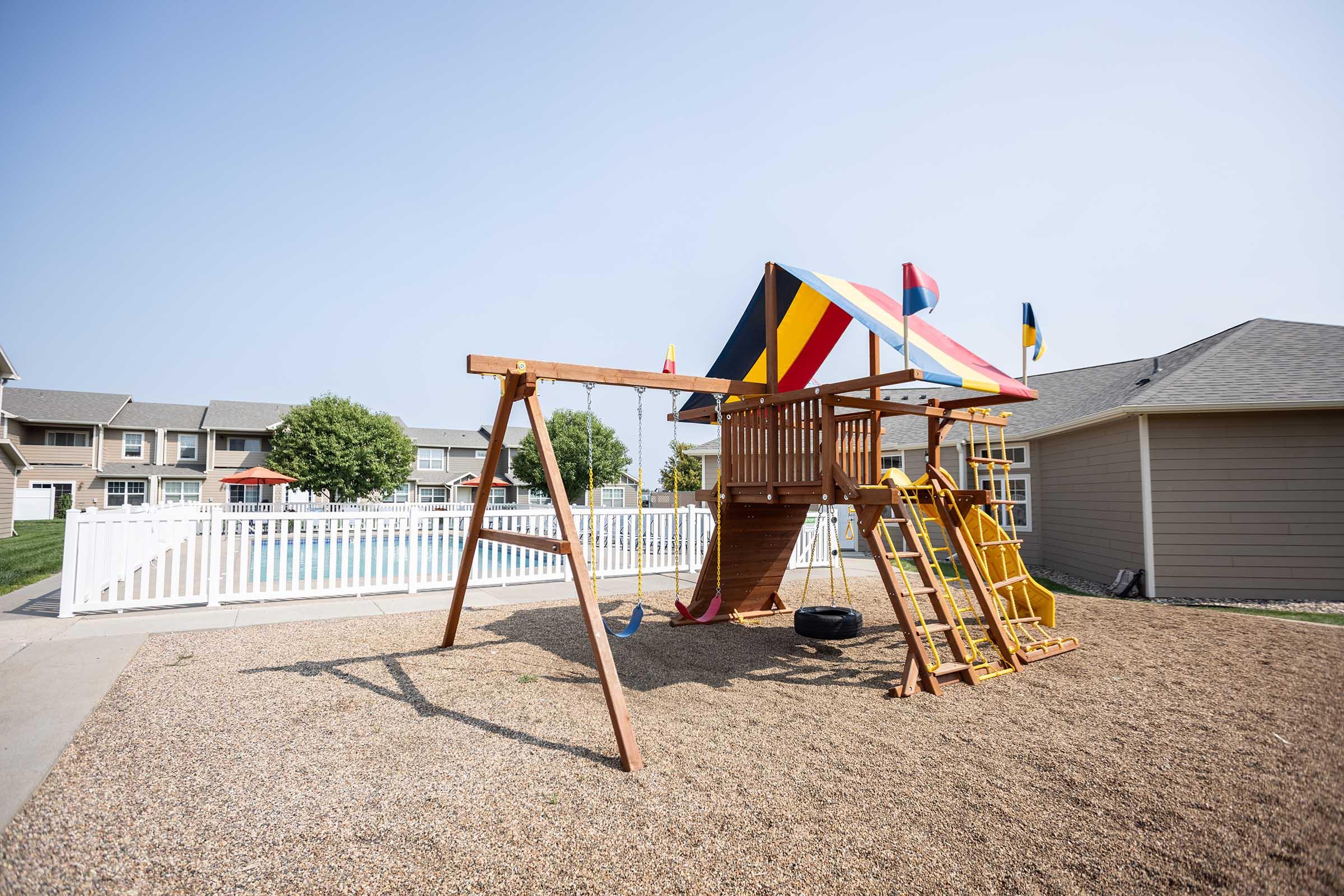 A colorful playground structure with a slide, swings, and a climbing wall situated in a gravel area. In the background, there is a swimming pool surrounded by a white fence and residential buildings. Clear blue sky above, creating a sunny and inviting atmosphere.