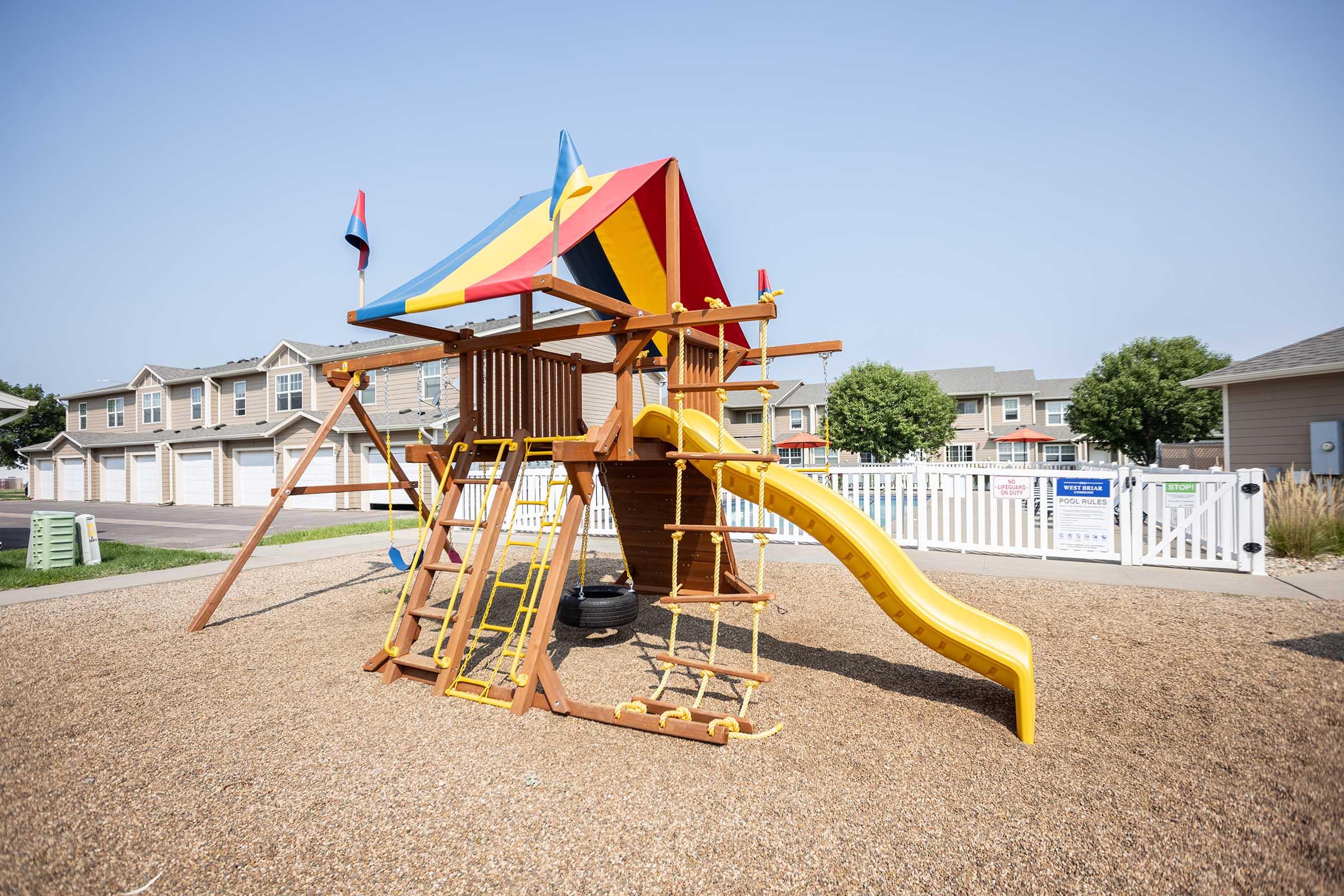 Colorful wooden playground structure with a slide, climbing ladder, and swings, set on a gravel surface. Brightly colored canopy on top, surrounded by a white fence and residential buildings in the background under a clear blue sky. Suitable for children's outdoor play.