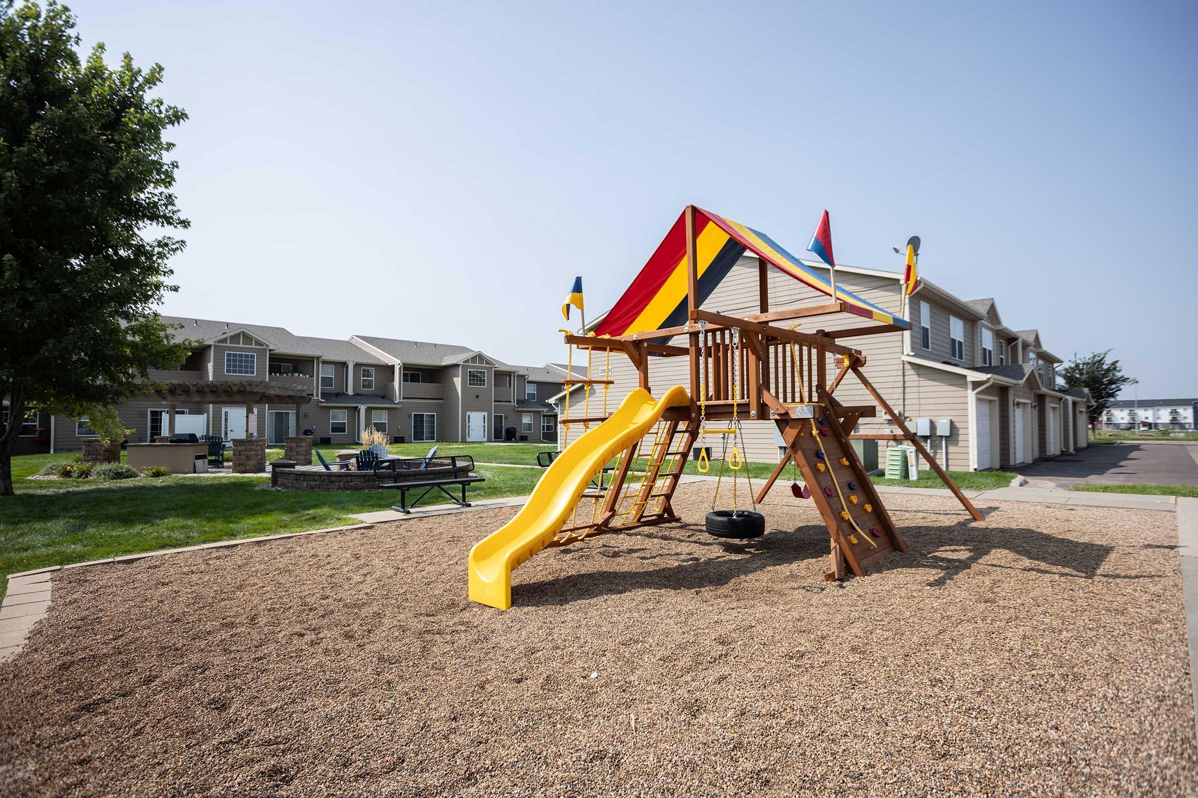 Playground structure with a colorful canopy, featuring a slide, swings, and a grassy area. In the background, there are residential buildings with a clear sky overhead. Gravel ground surrounds the play area, providing a safe surface for children to play.