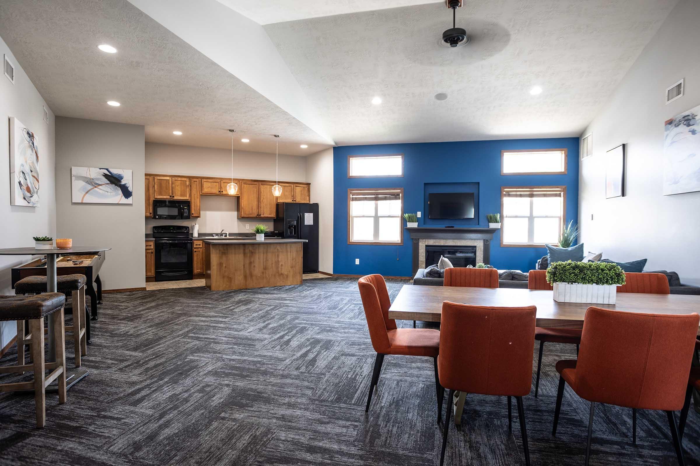 A modern, open-concept living space featuring a kitchen with wooden cabinets, a dining area with orange chairs, and a cozy seating area with a television. Large windows provide natural light, and decorative plants add a touch of greenery to the space. The flooring is dark, creating a stylish contrast.