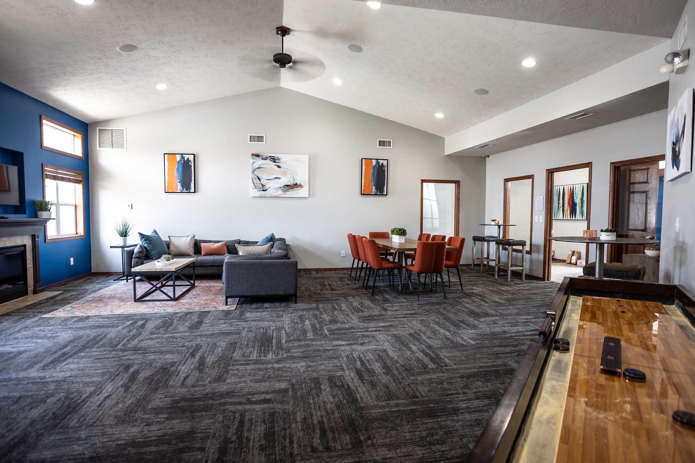 A spacious, modern living area featuring a gray sofa with orange accents, a round dining table with chairs, and a large window. The walls are painted in neutral tones with a blue accent wall. There is a coffee table, decorative plants, and a shuffleboard table on the side, creating a welcoming atmosphere.