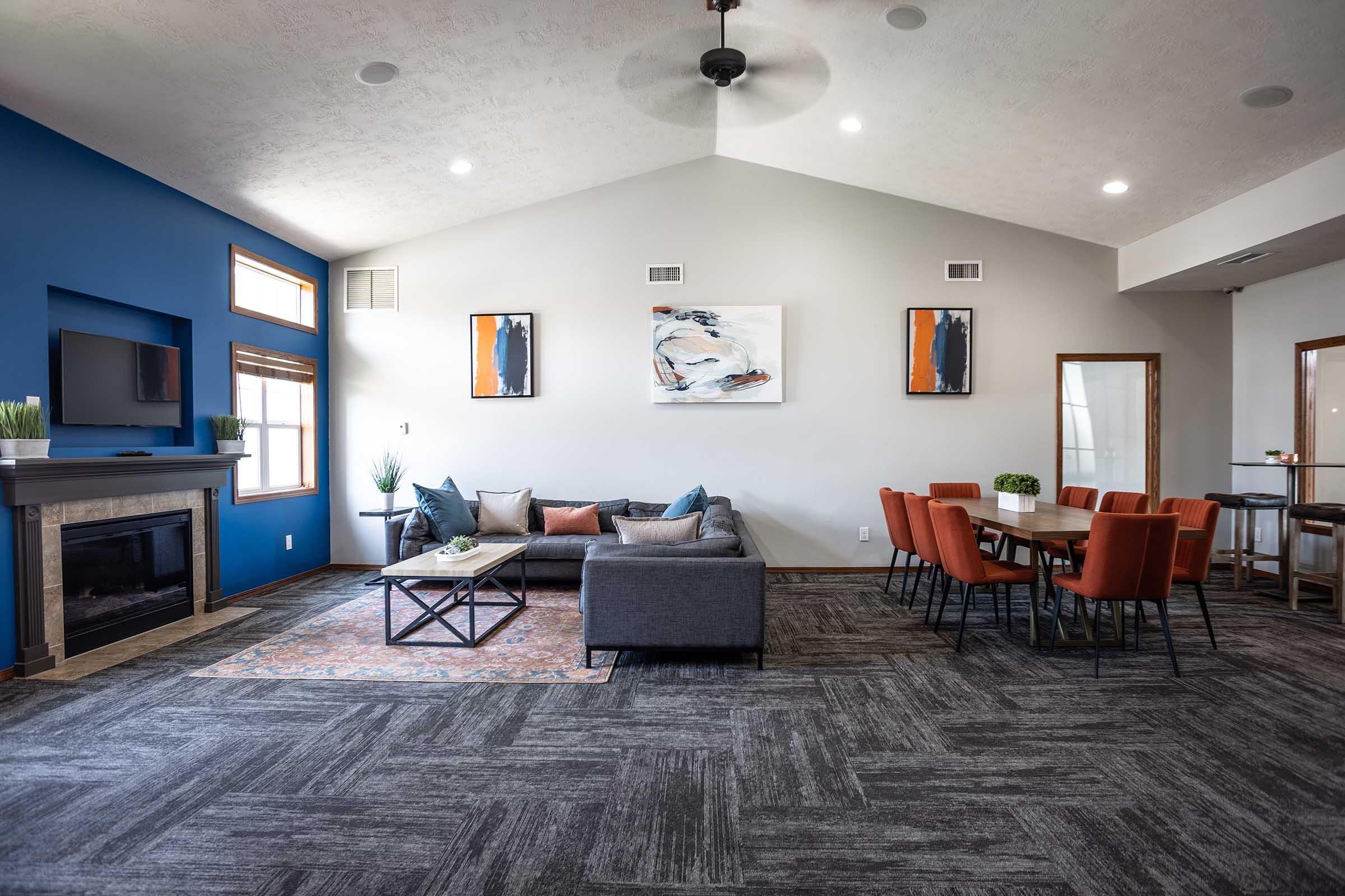 A modern living space featuring a gray sectional sofa with colorful pillows, a coffee table, and a flat-screen TV mounted on a blue accent wall. The room has a dining area with a long table surrounded by orange chairs. Natural light comes through large windows, enhancing the cozy atmosphere.
