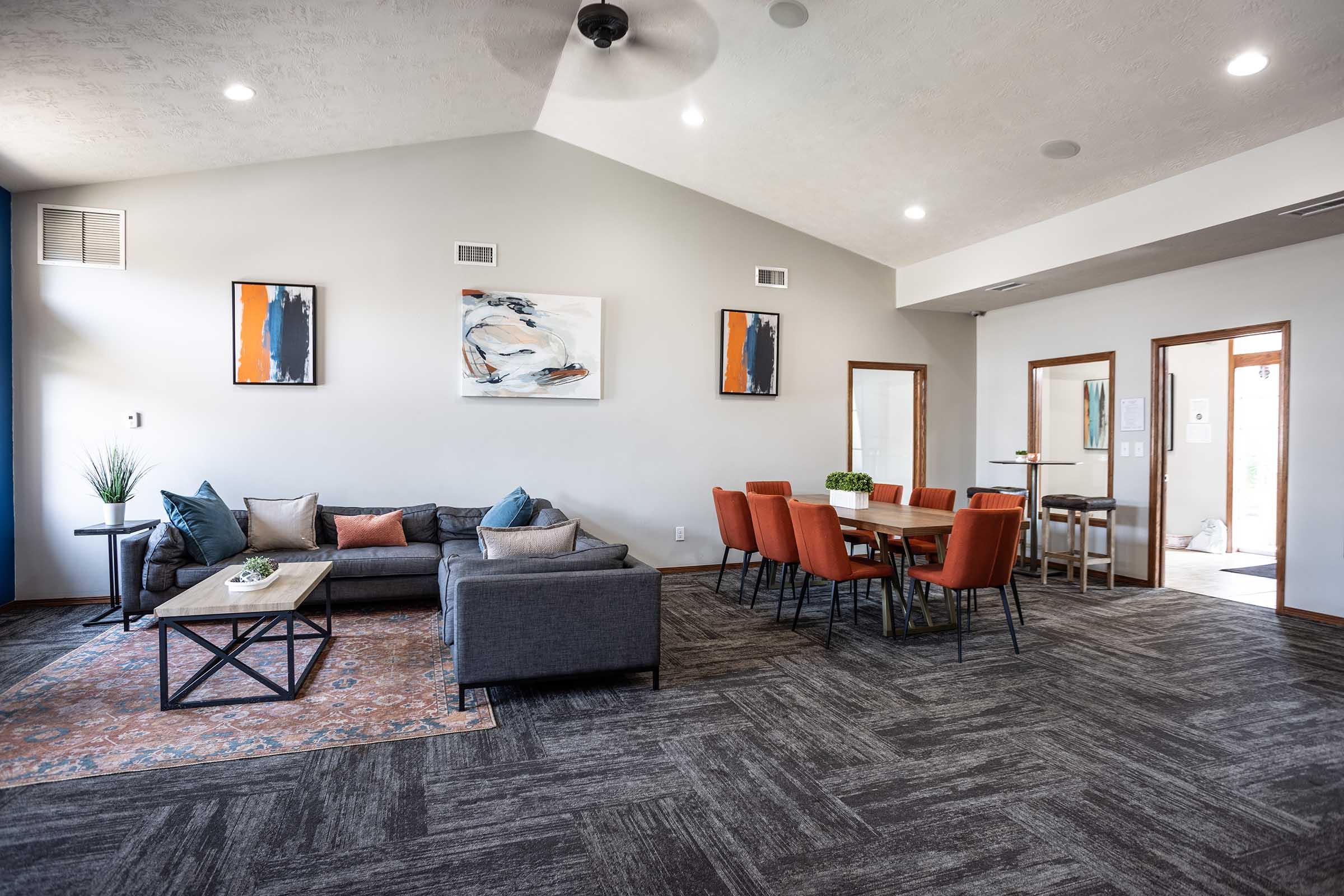 Modern living room with a gray sectional sofa, accent pillows, and a coffee table. Adjacent dining area features a wooden table with orange chairs. Walls are light gray with abstract artwork. Natural light enters through large windows, creating a bright and inviting atmosphere. Stylish decor adds a contemporary touch.