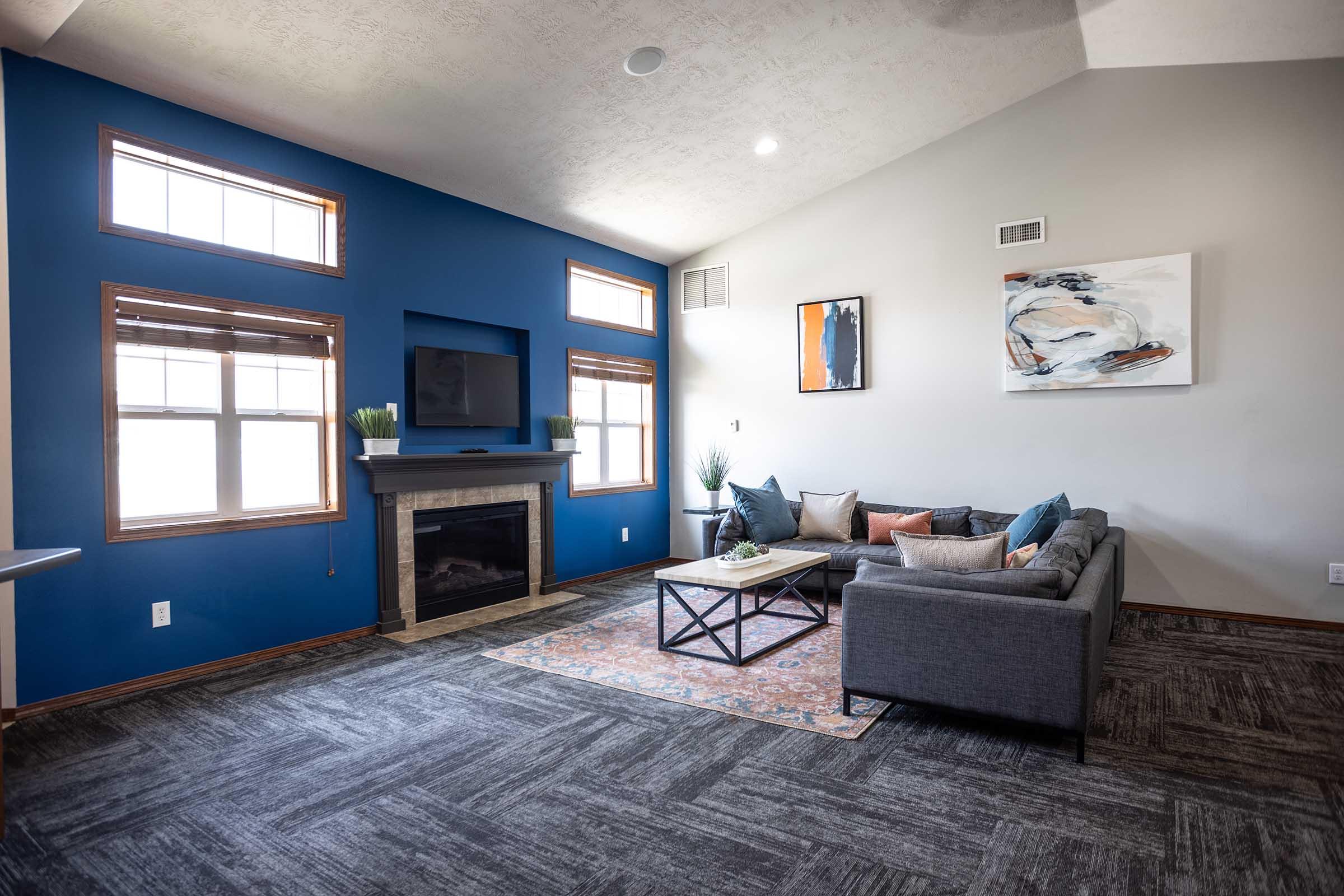 A bright living room featuring a blue accent wall, large windows, and a modern fireplace. There's a gray sectional sofa with decorative pillows, a coffee table, and an area rug. A flat-screen TV is mounted on the wall, and the space is decorated with plants and artwork, creating a stylish and inviting atmosphere.