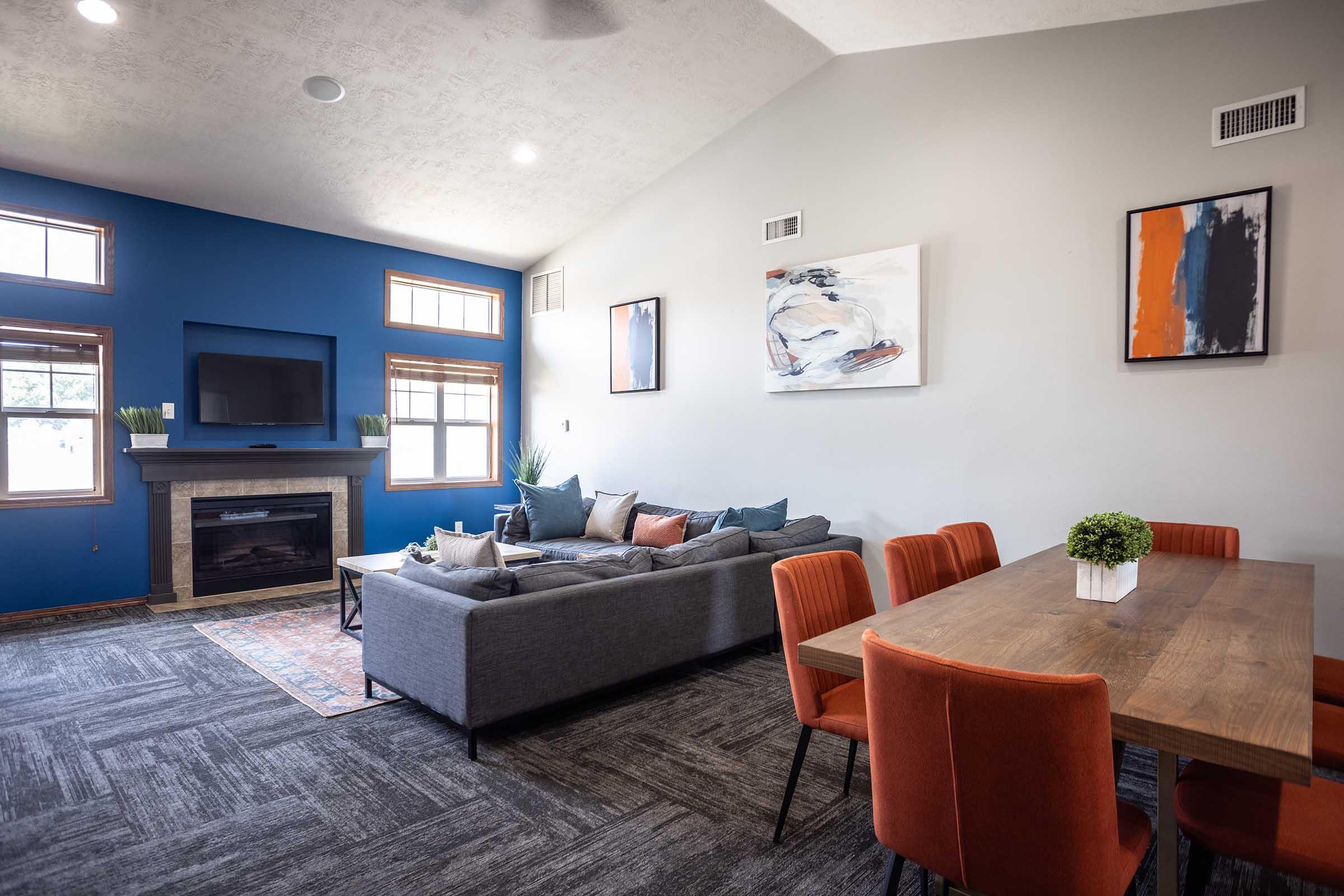 A modern living area featuring a gray sectional sofa, a wooden dining table with orange chairs, and a blue accent wall. There is a television mounted on the wall, a fireplace below it, and artwork framed on the walls. The space is well-lit with large windows, creating a cozy atmosphere.