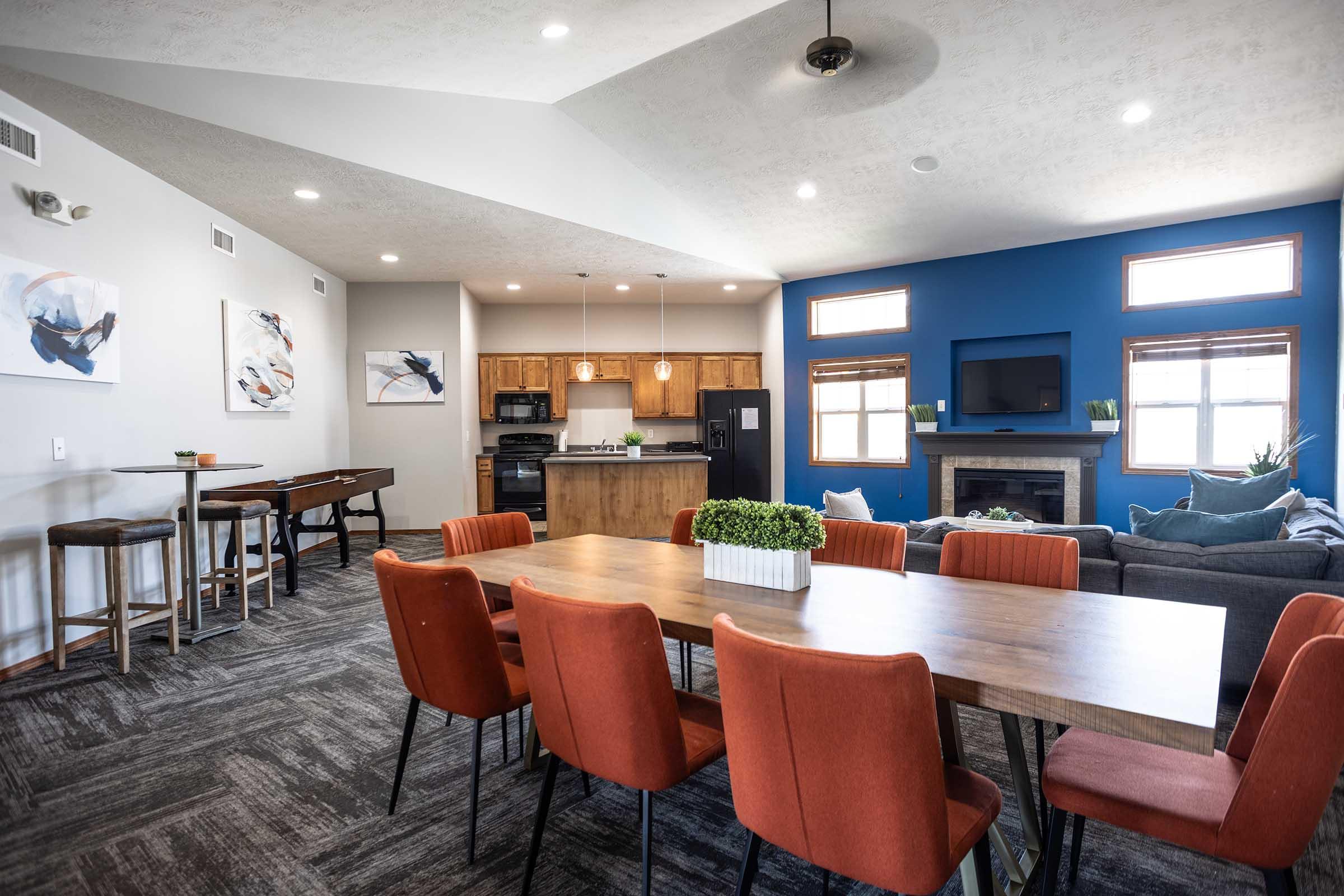 Interior of a modern open-plan living area featuring a dining table with orange chairs, a cozy seating area with a gray sofa, and a kitchen with wooden cabinets. The room has a blue accent wall and large windows, creating a bright, inviting atmosphere. Art pieces are displayed on the walls.