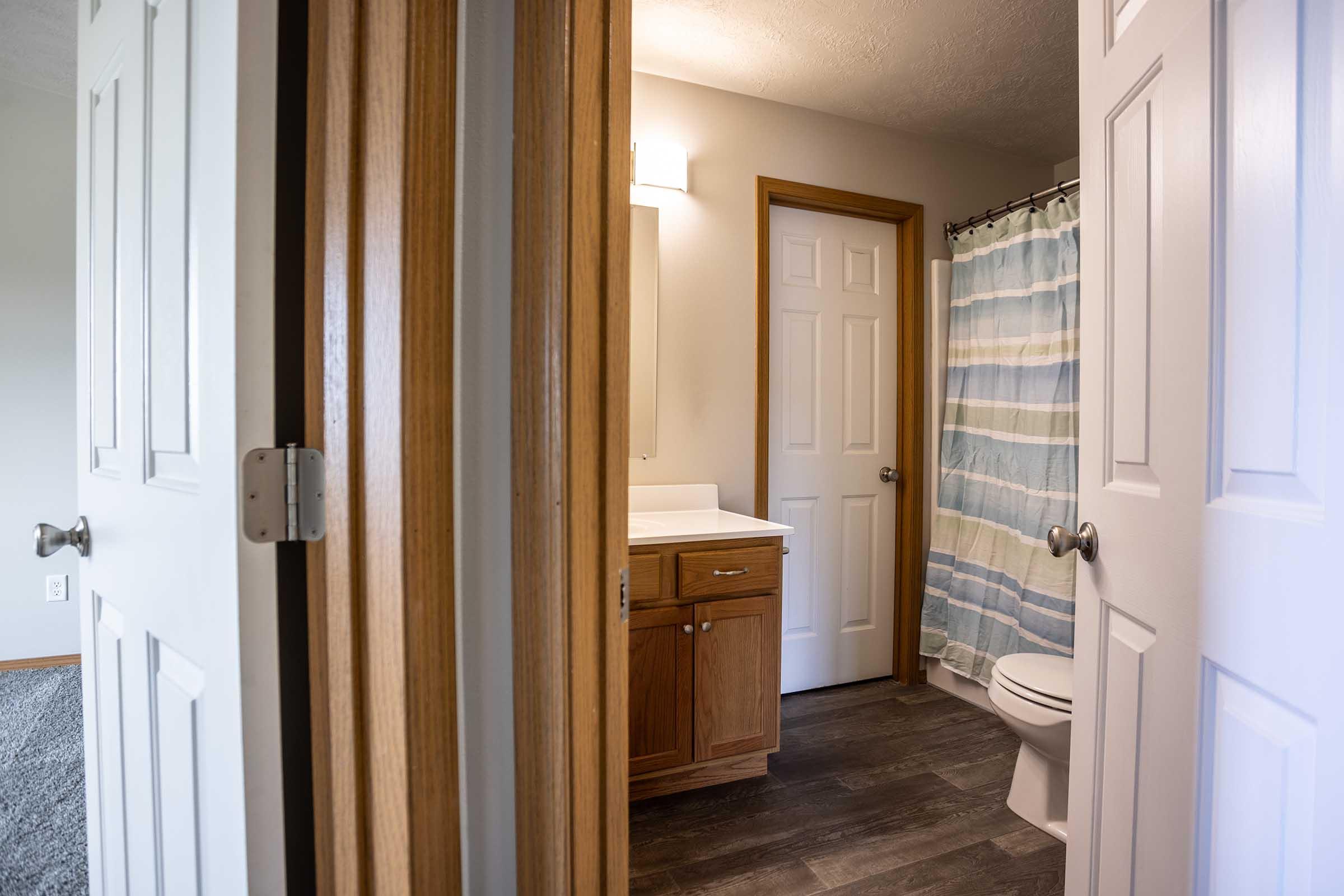 A view of a bathroom from an adjoining room, featuring a white vanity with a mirror, a shower curtain with blue and white stripes, and a toilet. The doors to the bathroom and adjoining room are partially open, revealing light-colored walls and a light fixture. Hardwood or laminate flooring is visible.