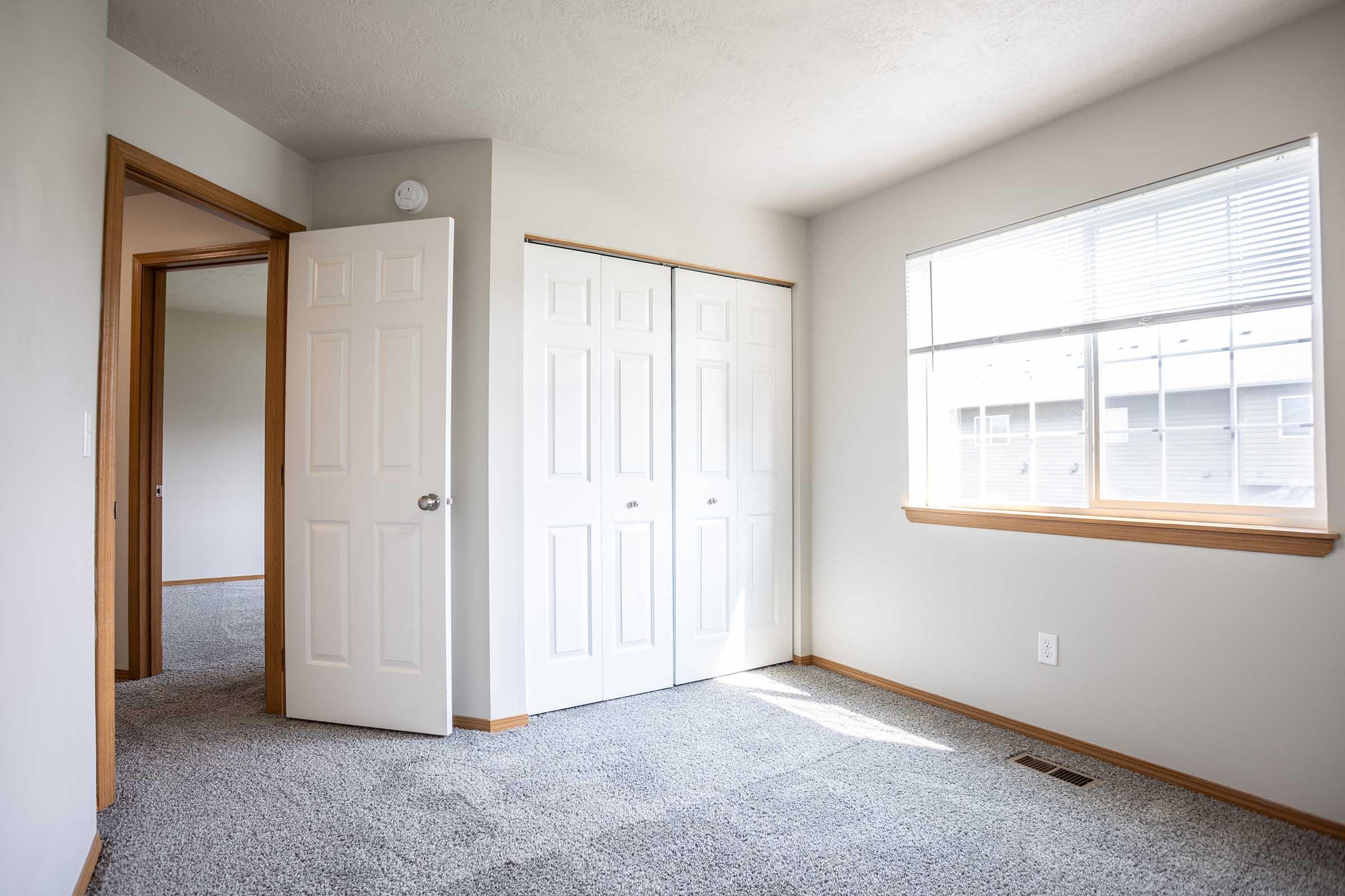A well-lit, empty room featuring beige walls and carpet. There is a window with natural light and a view outside. To the left, a doorway leads to another room, and there are double white closet doors on the right. The space appears tidy and freshly painted, ideal for moving in or decorating.