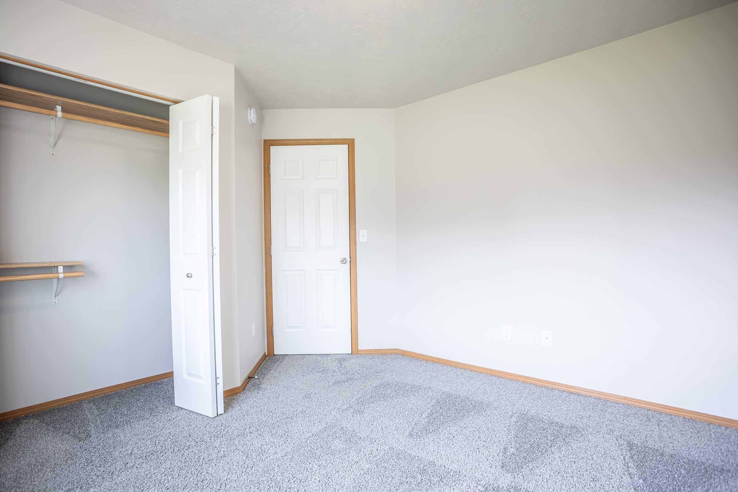 A blank, unfurnished room with light gray walls and carpet. There is a closed closet with a shelf on the left and a white door leading to another space on the right. The overall lighting is bright and the atmosphere is clean and simple.