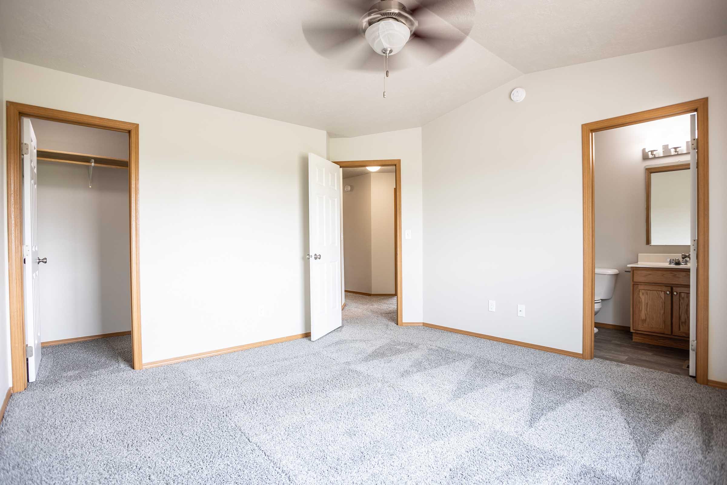 A spacious, empty room with light gray carpet. It features two doorways leading to adjacent areas: one to a closet and another to a bathroom. A ceiling fan is visible, and the walls are painted a neutral color, creating a bright and inviting atmosphere.