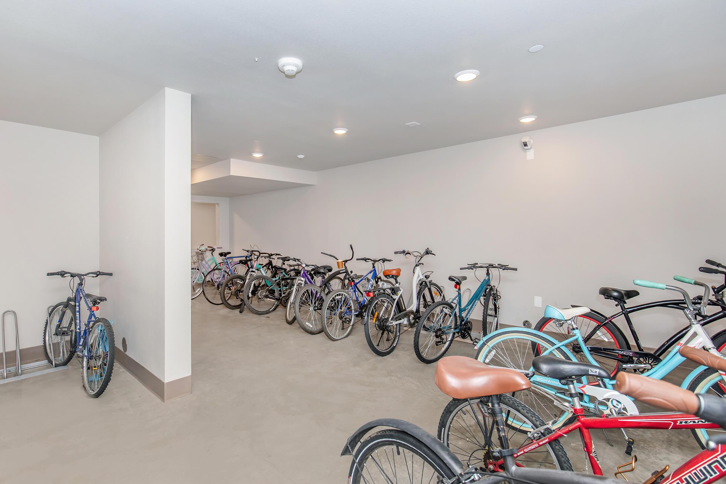 A well-lit indoor bicycle storage area with several bikes parked in designated spaces. The floor is concrete, and there are wall-mounted bike racks. The space is clean and organized, providing ample room for multiple bicycles.