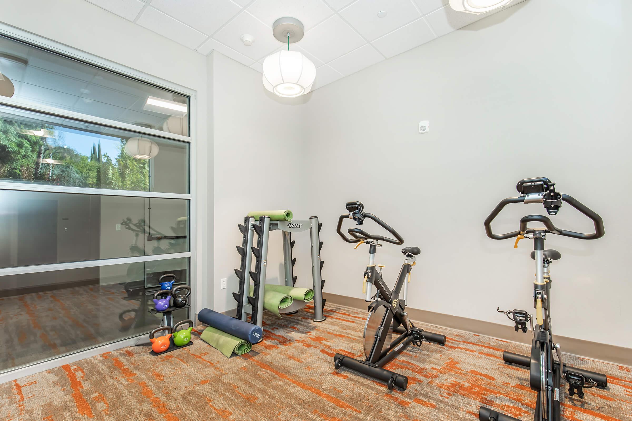 A well-lit fitness room featuring two stationary bikes, a rack with kettlebells, and rolled yoga mats on the floor. The modern space includes large windows and contemporary lighting fixtures, creating an inviting atmosphere for workouts.