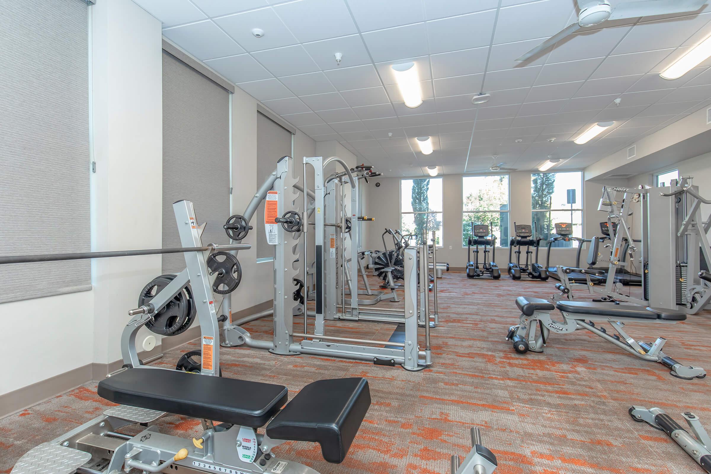 A modern fitness center featuring various exercise equipment including weight machines, cardio machines, and free weights. The space has large windows allowing natural light, with a clean and organized layout, suitable for workouts. The flooring is carpeted with a bright orange and gray pattern.