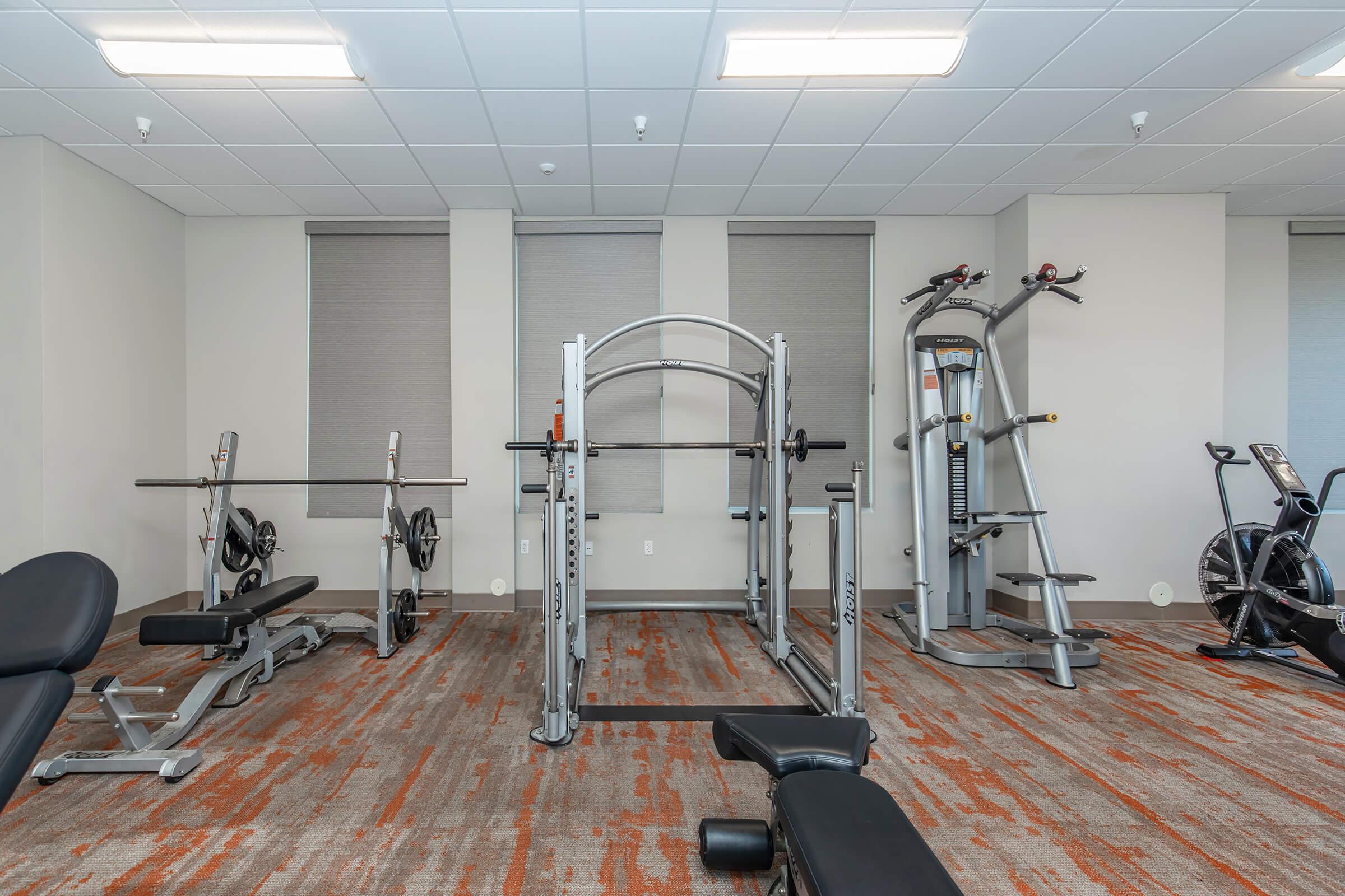 A modern gym interior featuring various fitness equipment. There is a weight bench, a barbell rack with weight plates, a multi-functional workout station, and a stationary bike, all set against a well-lit background with two windows covered by shades. The flooring is carpeted in warm tones.