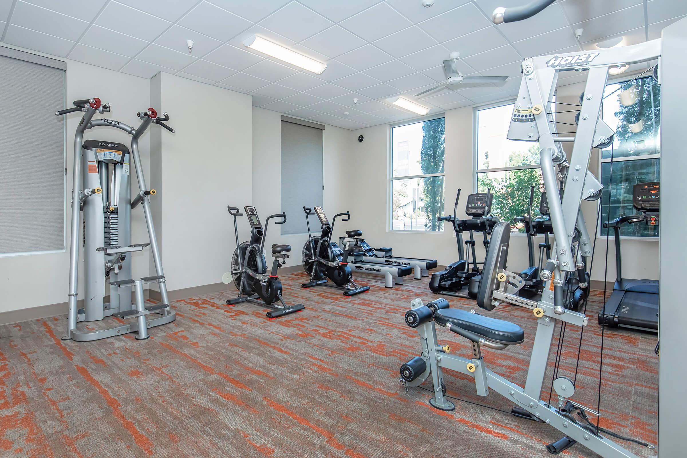 A modern fitness room featuring various exercise equipment, including stationary bikes, an elliptical machine, a weight training machine, and a treadmill, all arranged in a well-lit space with large windows and a carpeted floor.